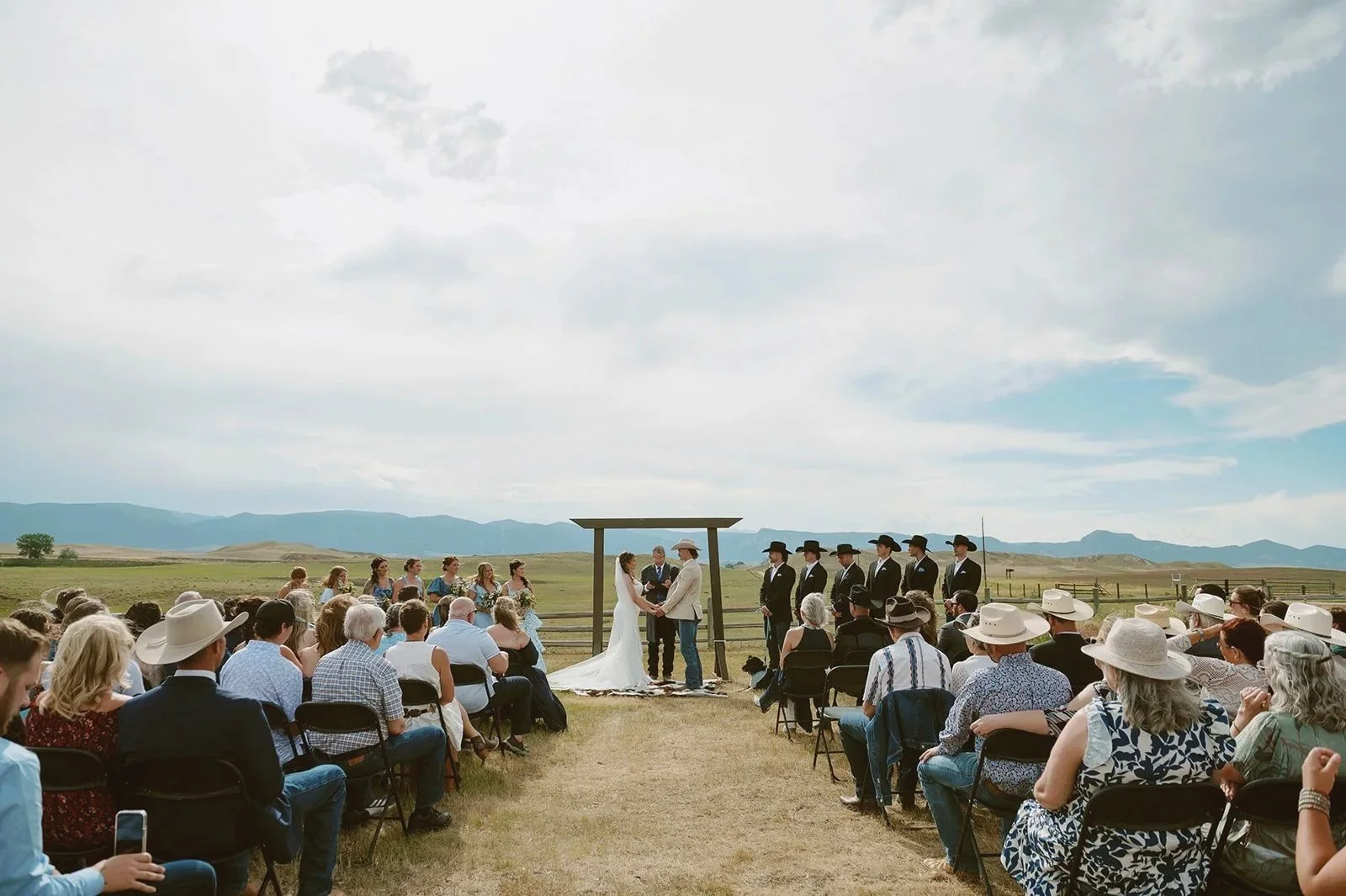 Outdoor wedding ceremony with a bride and groom standing under a wooden arch, surrounded by seated guests in a rural landscape with mountains in the background.
