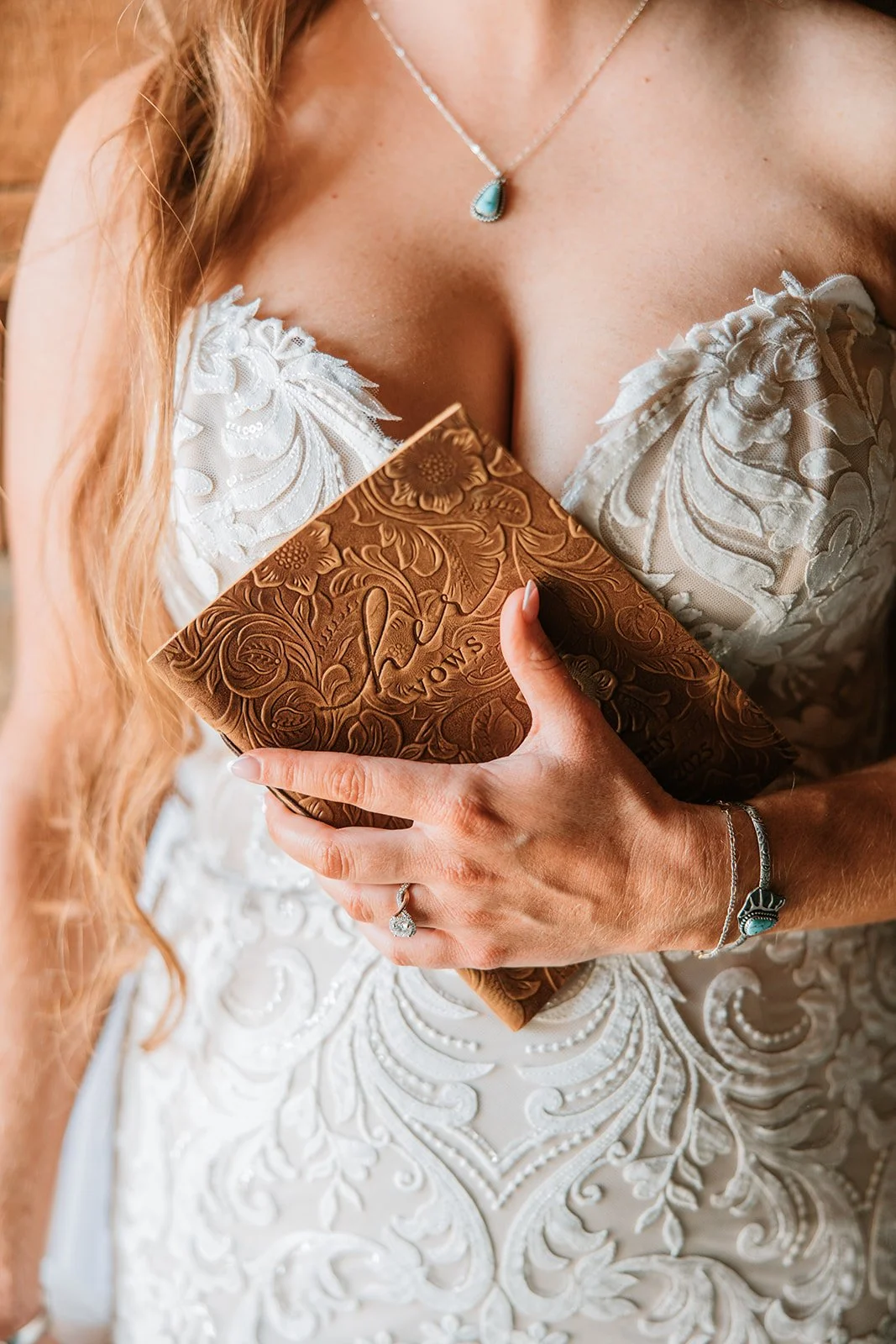 A bride holding a brown leather book or box with floral embossing, wearing a white lace wedding dress with intricate embroidery, a silver necklace with a turquoise pendant, a silver bracelet, and a ring with a large gemstone.