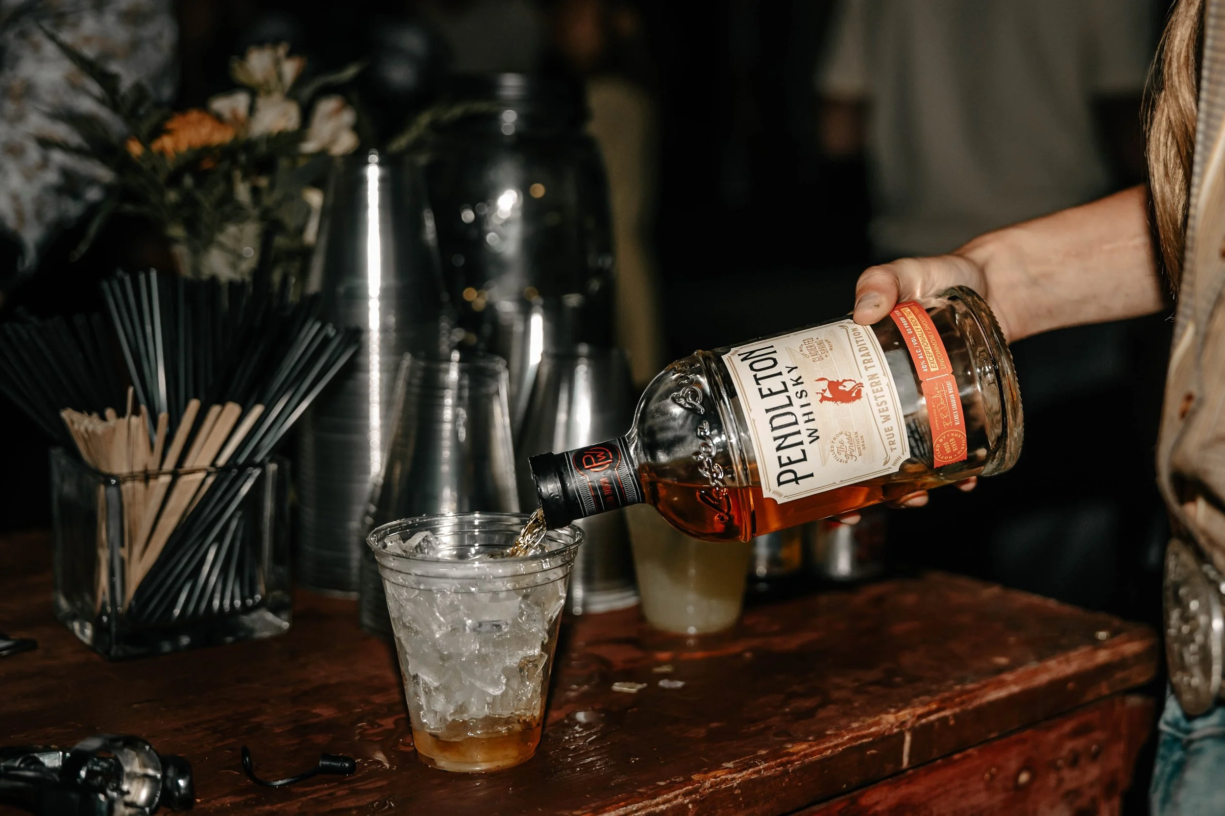 A person pours Pendleton whisky over ice in a glass on a bar counter, with bar tools and straws visible in the background.