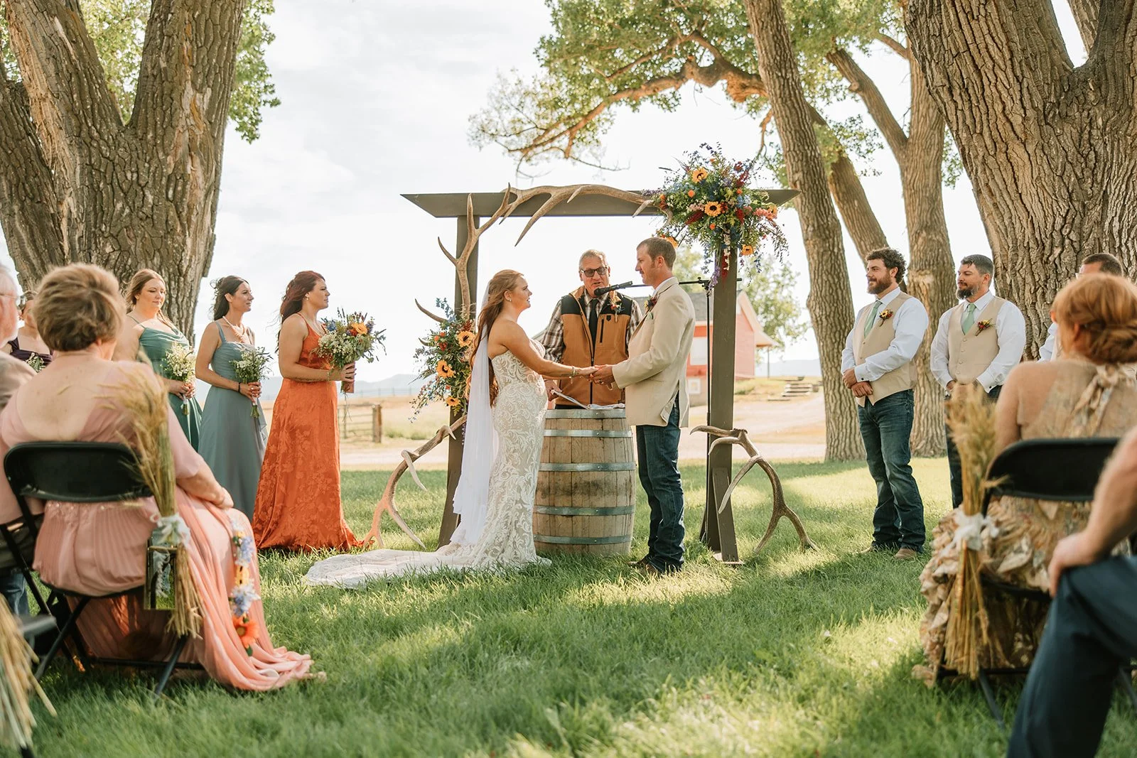 A couple exchanging vows at an outdoor rustic wedding ceremony under large trees, with bridesmaids and groomsmen standing nearby, and guests seated watching.