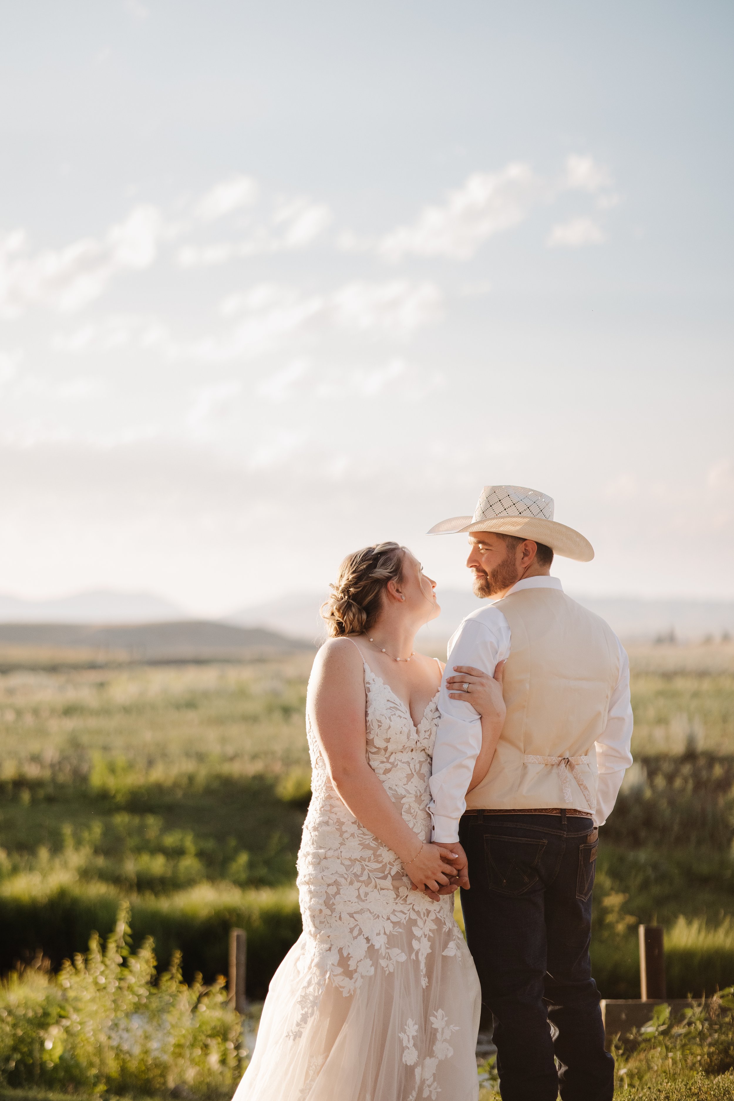 A bride and groom holding hands and gazing at each other outdoors during sunset, with a scenic landscape and cloudy sky in the background.