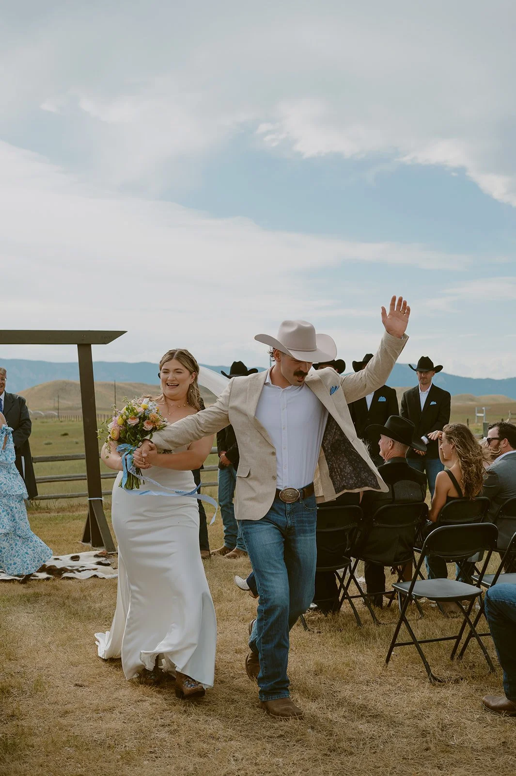 A bride and groom walking hand in hand during an outdoor wedding ceremony, with guests seated on chairs and a pastoral landscape in the background.