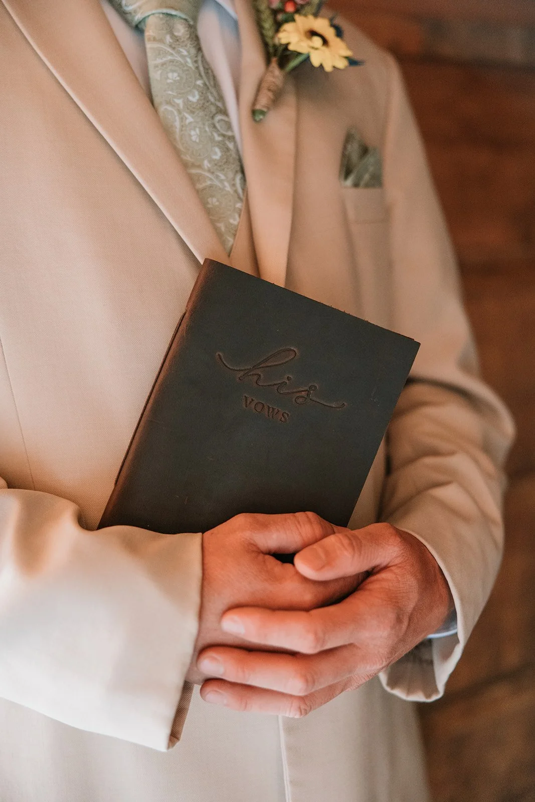 A person dressed in a beige suit holding a black book titled 'His Vows'. The person also has a boutonniere with yellow flowers on their suit lapel.