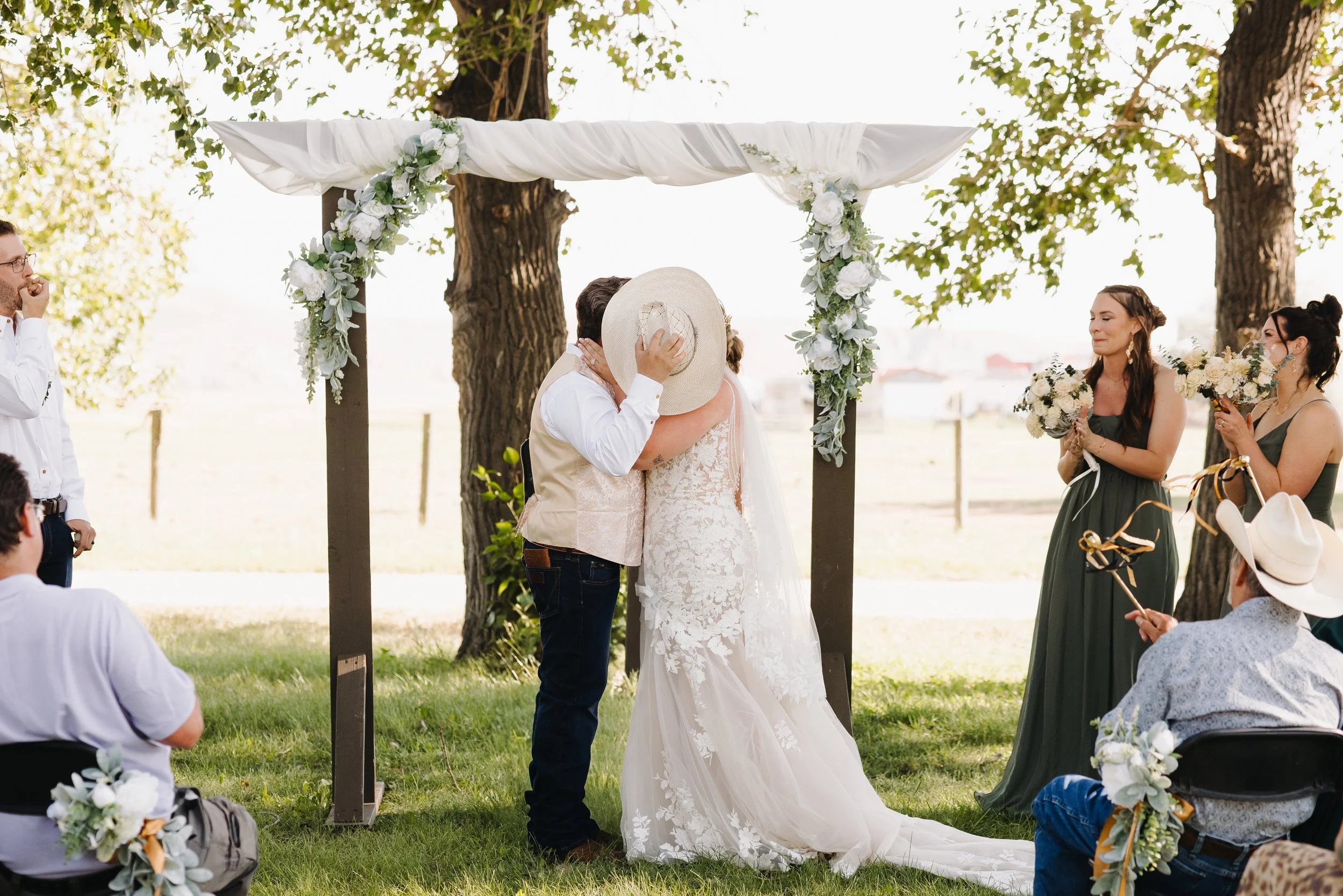 A couple shares a kiss during an outdoor wedding ceremony, with the officiant holding a large sun hat to cover their faces, under a floral arch between two large trees. Bridesmaids are holding bouquets, and guests are seated on grass with floral decorations.