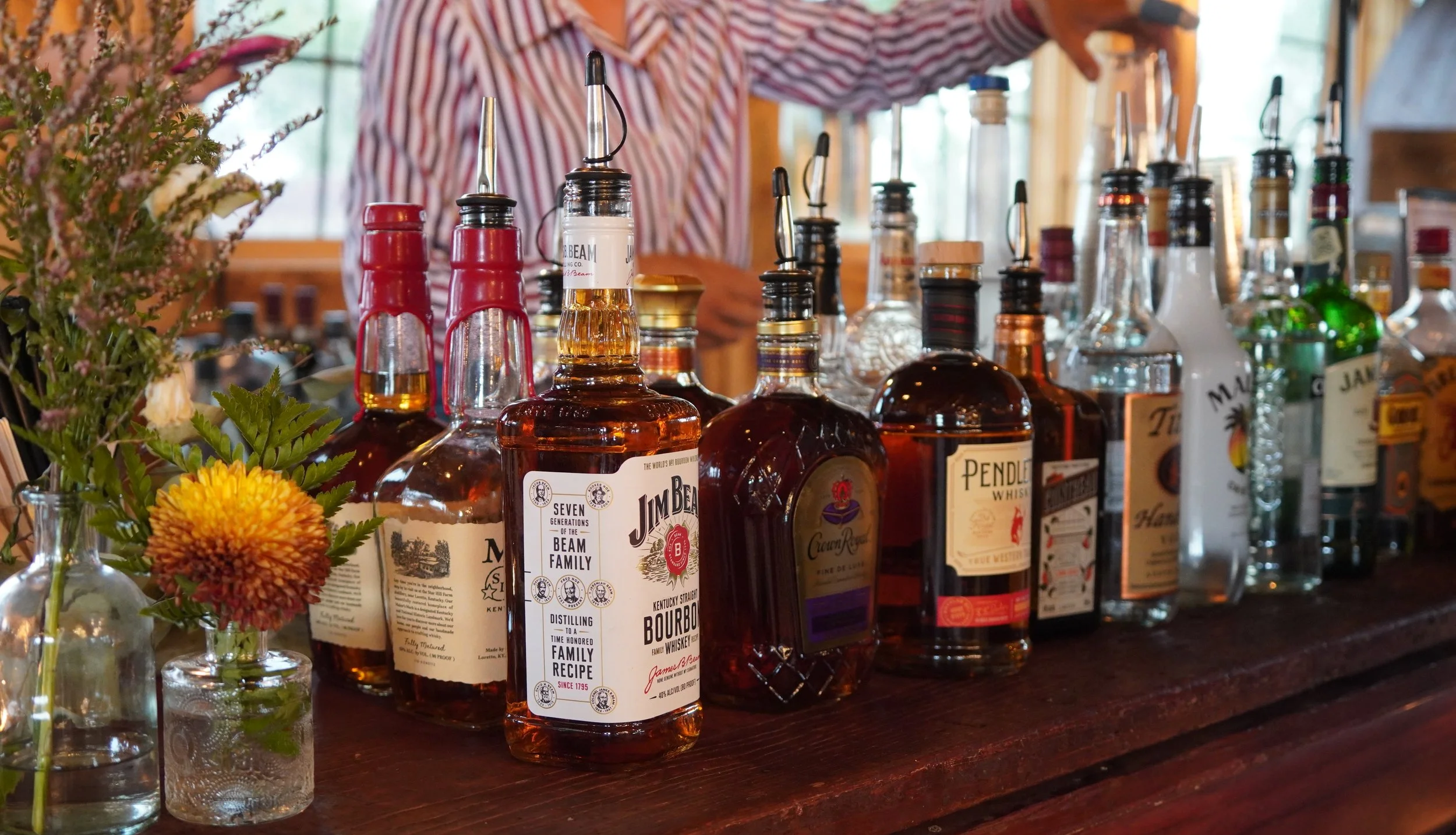A shelf stocked with various bottles of alcohol including bourbon and whiskey, with a bartender in the background and flowers in vases on the left.