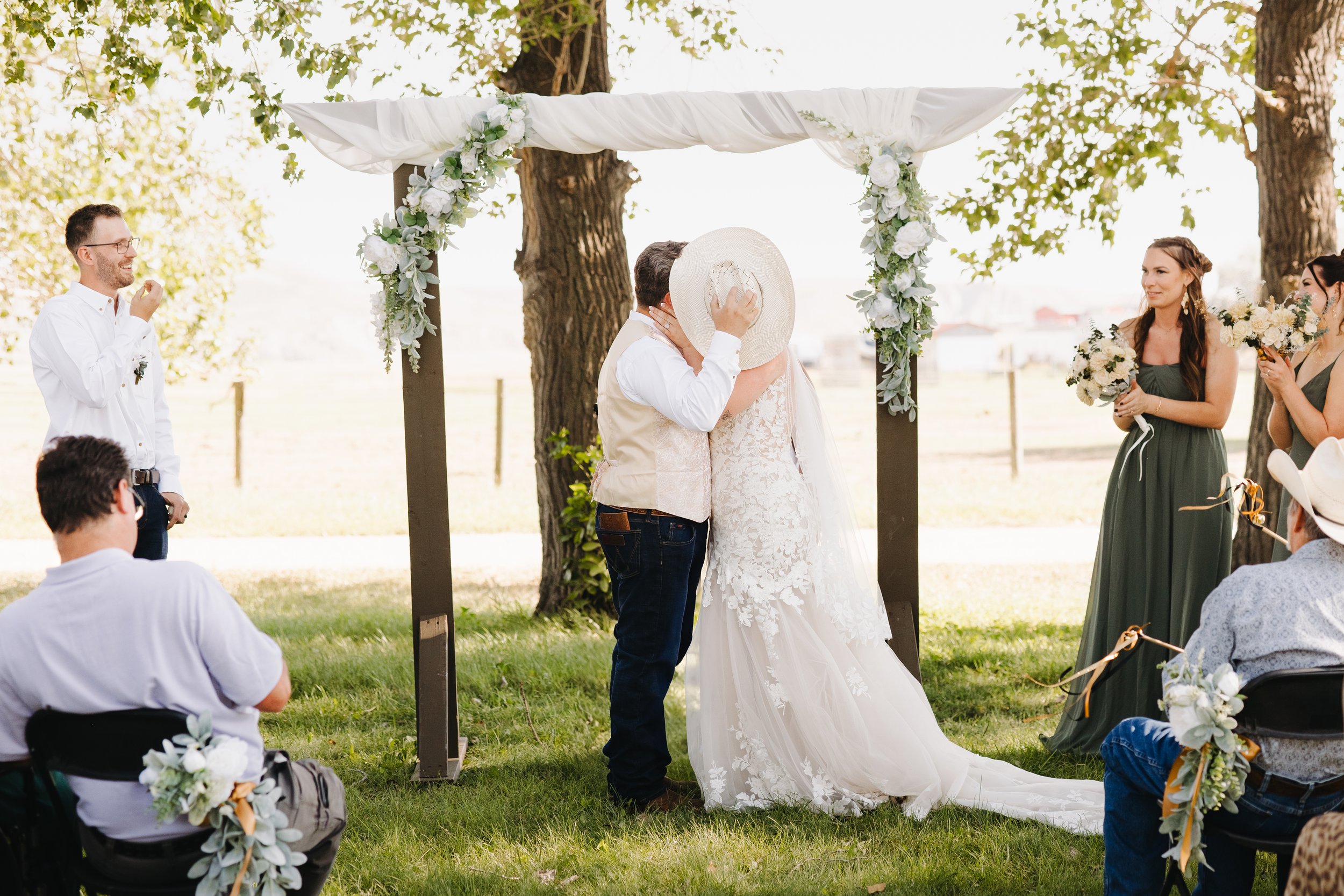 Couple kissing during outdoor wedding ceremony under a decorated arch with pavilion, surrounded by guests and bridesmaids, in a park-like setting.