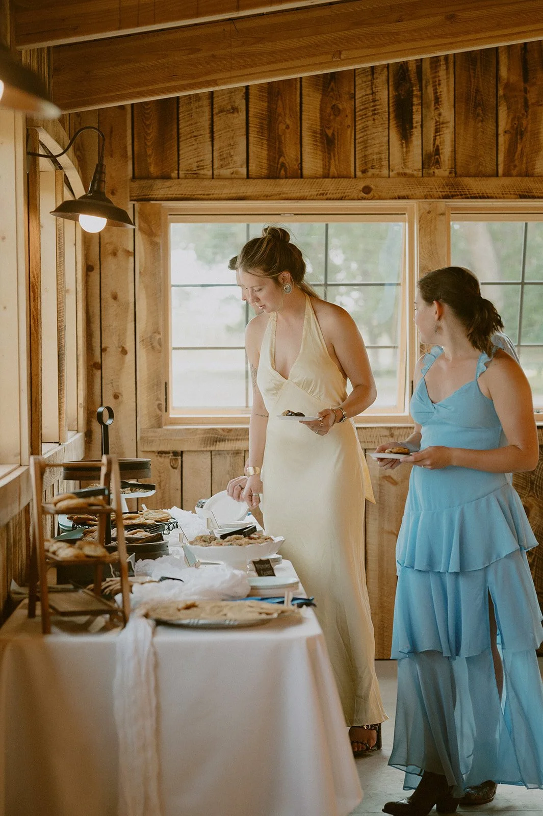 Two women in elegant dresses standing at a dessert table in a rustic wooden room with large windows, serving themselves sweet treats during a daytime celebration.