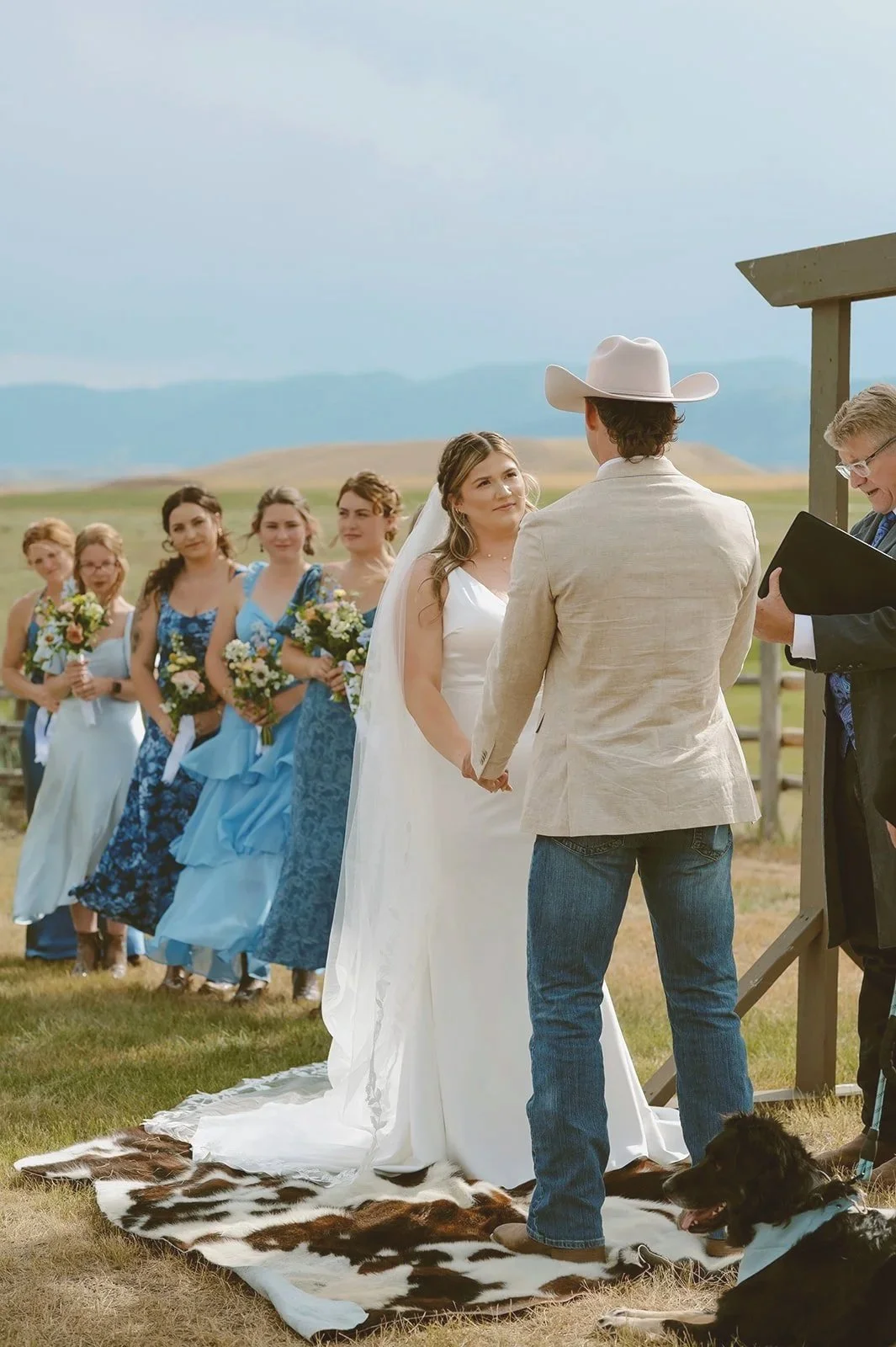 A wedding ceremony outdoors with a bride and groom holding hands, standing on a cowhide rug, with a officiant reading. Bridesmaids in blue dresses and holding bouquets stand behind them. The setting features open fields and mountains in the background.