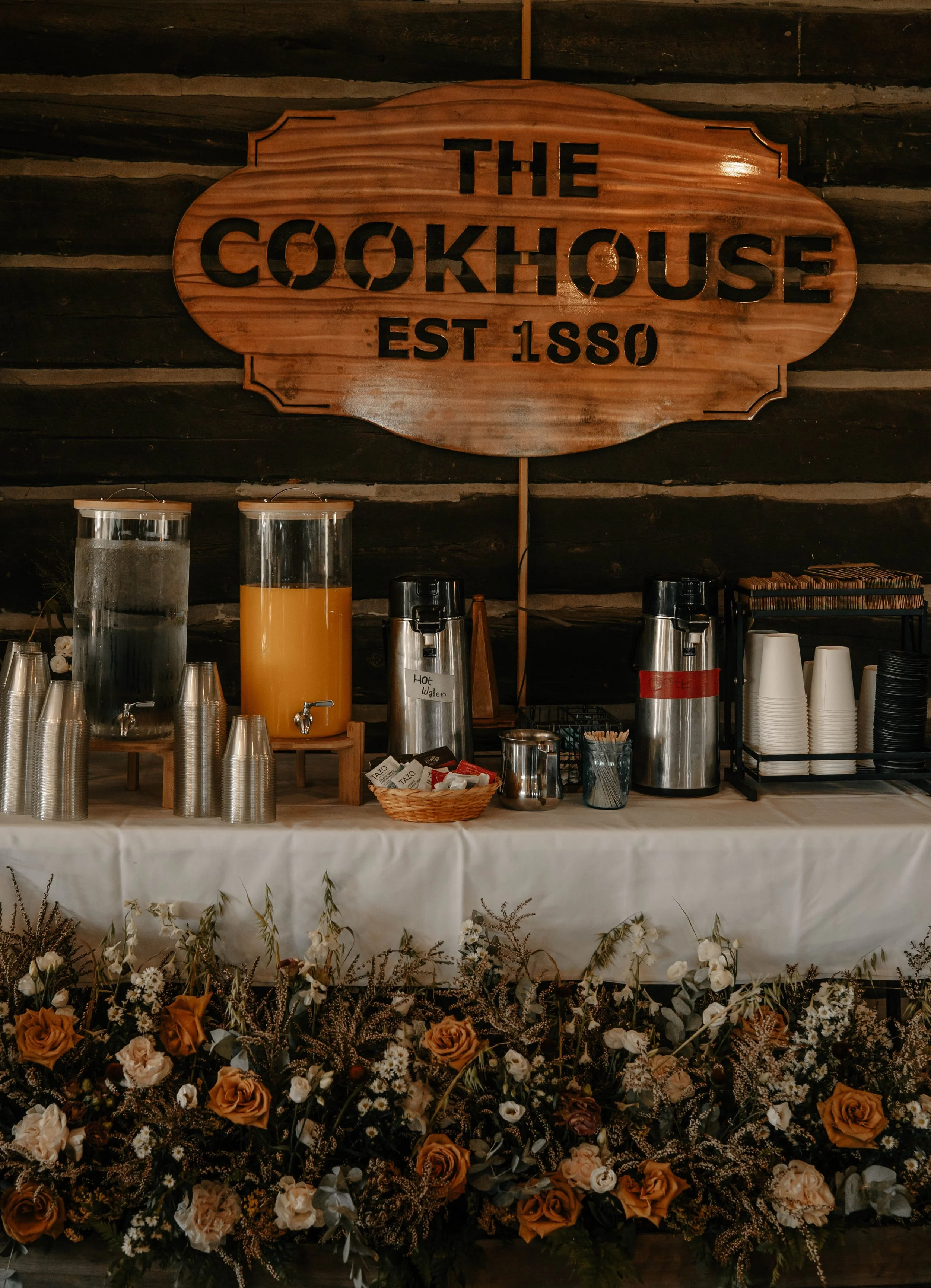 A beverage station setup with two large glass dispensers, one containing water and the other orange juice, on a white tablecloth. There are stacks of disposable cups, stir sticks, and a basket of tea bags. A large wooden sign above reads 'The Cookhouse Est 1880.' The table is decorated with a floral arrangement of roses and greenery in front, against a wooden wall background.