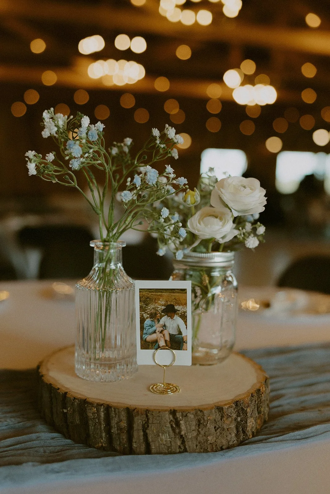 A table centerpiece with two glass jars holding white and blue flowers, a small framed photo of two people sitting outdoors, all displayed on a wooden round slice at a decorated event