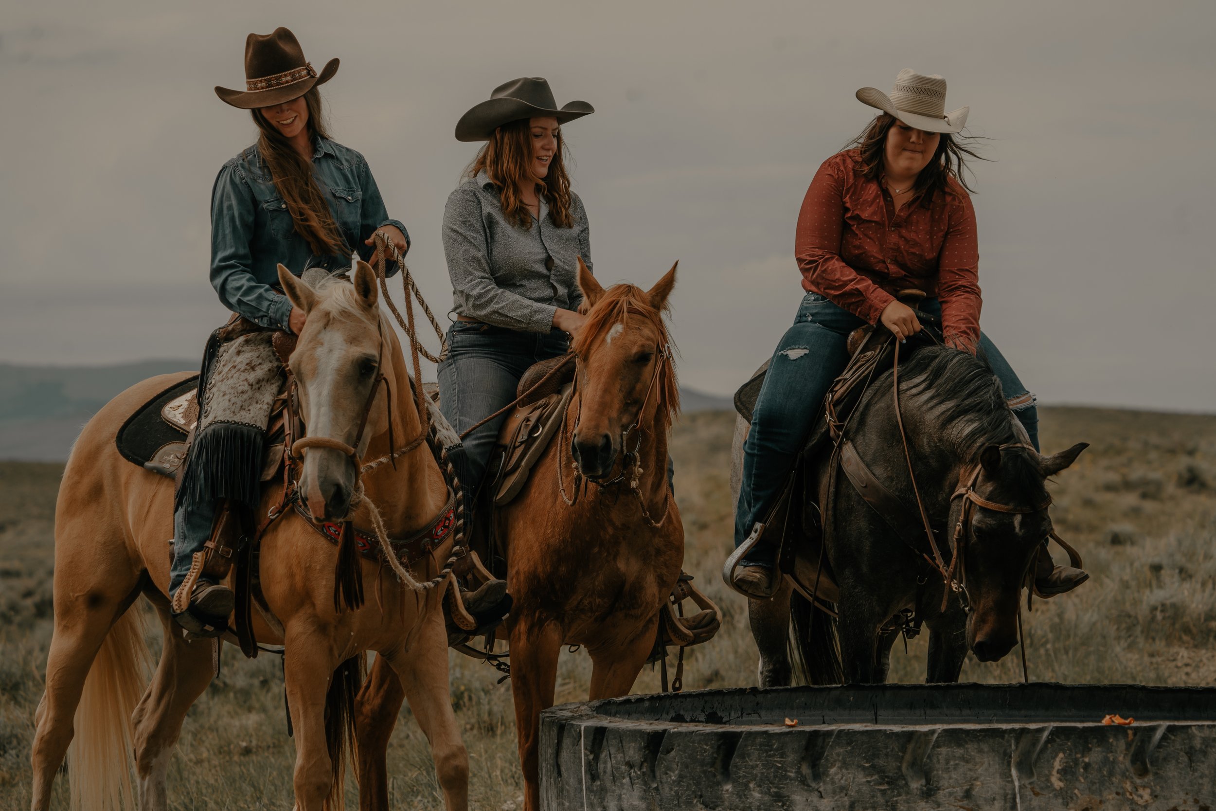 Three women riding horses outdoors in a rural landscape, all wearing cowboy hats and casual western attire.