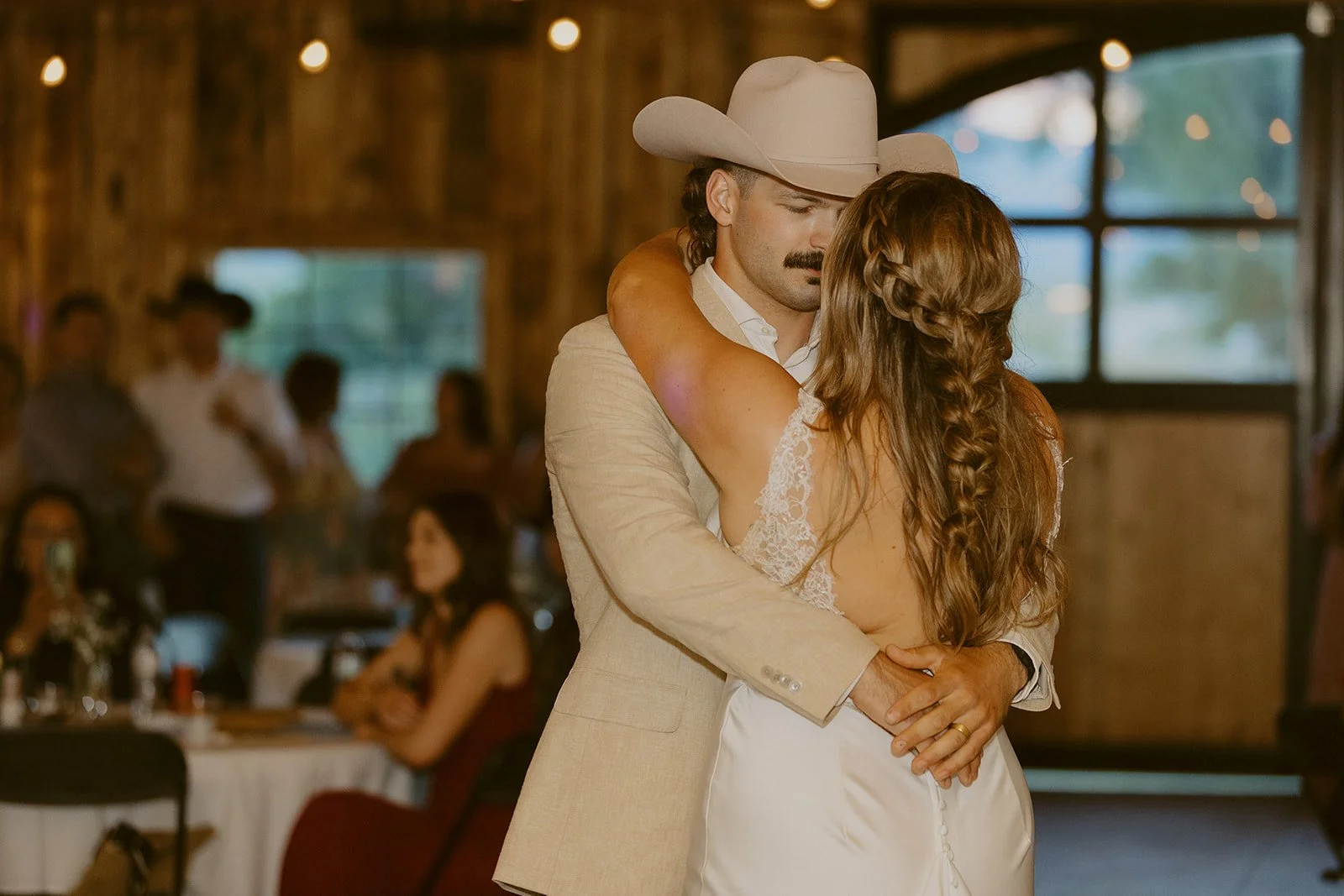 A man in a beige suit and cowboy hat dancing with a woman in a white lace dress during a wedding reception in a rustic barn.