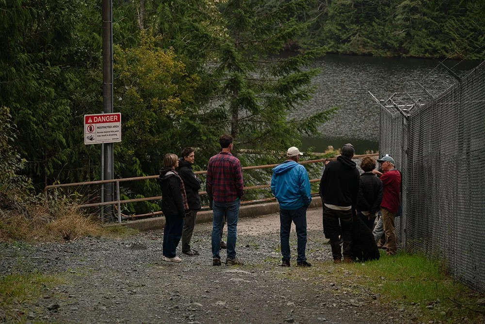 A group of people next to a chainlink fence with a water reservoir in the background.