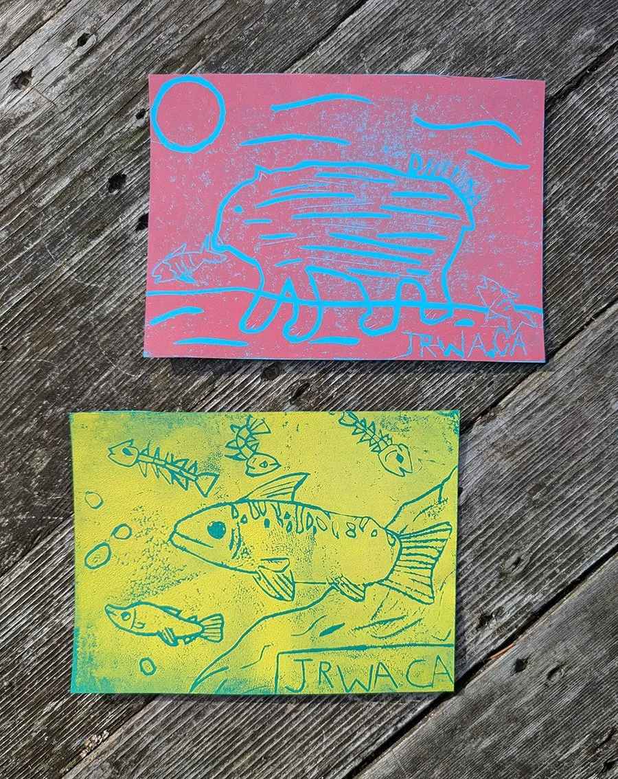2 pieces of cildrens artwork against a wood background, blue line work depicting a bear against a red background and blue line work depicting salmon against a yellow background 