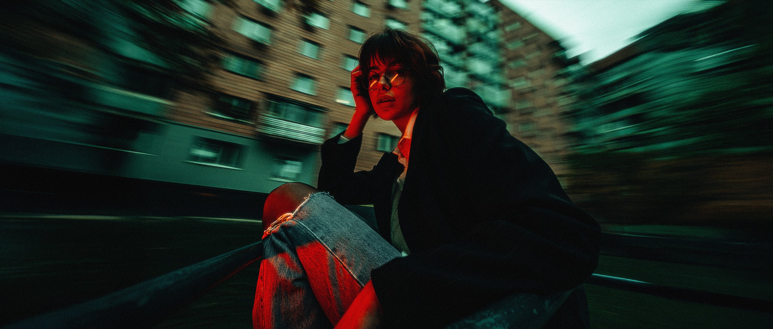 Young woman with glasses and short hair crouching on a rooftop or ledge in an urban area at night, with blurred buildings in the background, illuminated by red and greenish lighting.