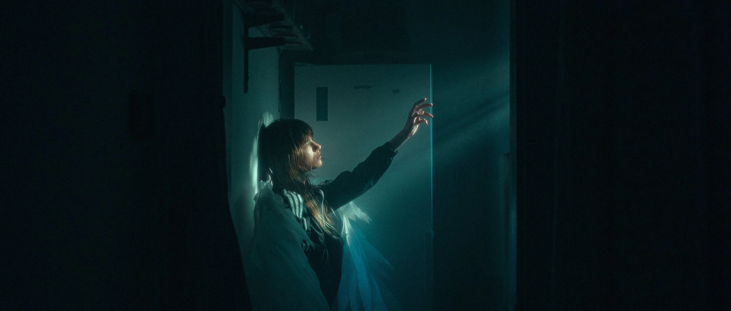 A person with long hair sitting in a dimly lit room, reaching out towards a light shining through a door, with sunlight streaming in from the right side.