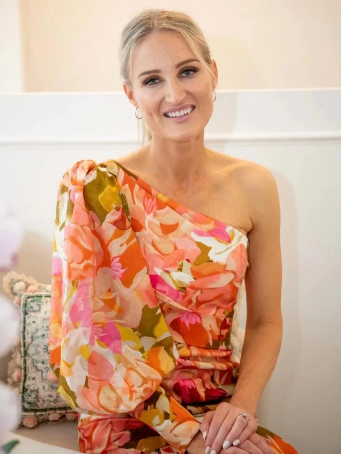 A woman with blonde hair, wearing a floral print dress with pink, orange, white, and green colors, sitting on a beige bench with a floral pillow in the background, smiling at the camera.