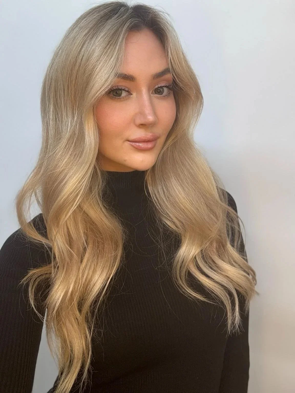 A woman with long, wavy blonde hair, wearing a black top, posing against a plain white background.