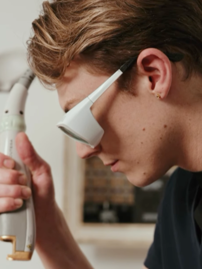 A person wearing magnifying glasses closely examining a sample under a microscope in a lab.