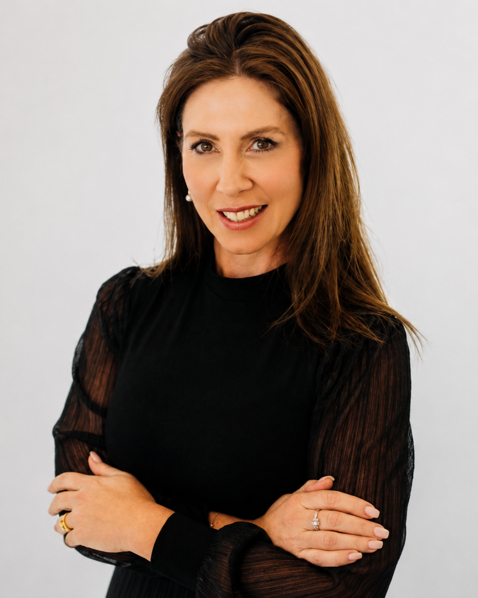 Portrait of a woman with brown hair wearing a black top with sheer sleeves, smiling with arms crossed against a plain light background.