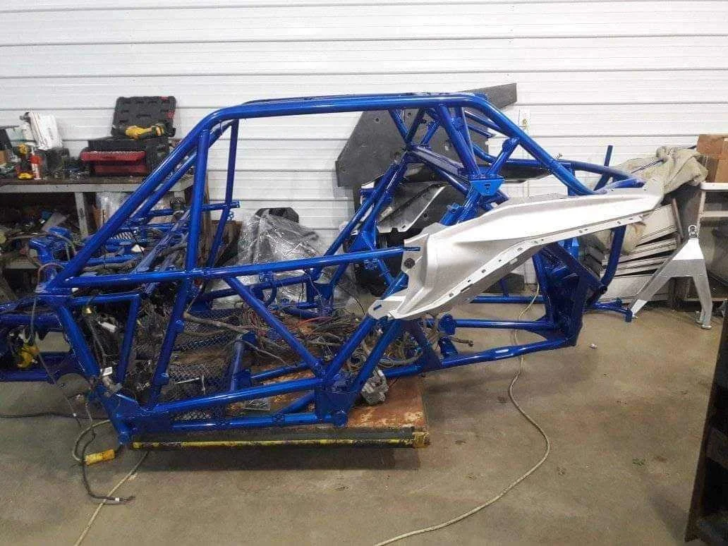 A blue car chassis frame with a white part on the side is placed on a wooden platform in a workshop.