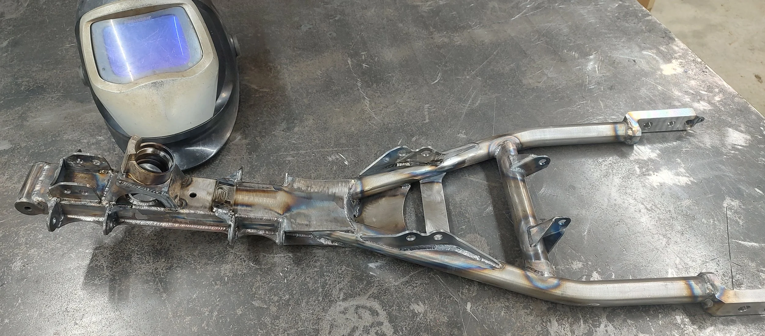 Metal motorcycle frame parts with a welding helmet on a workshop table.