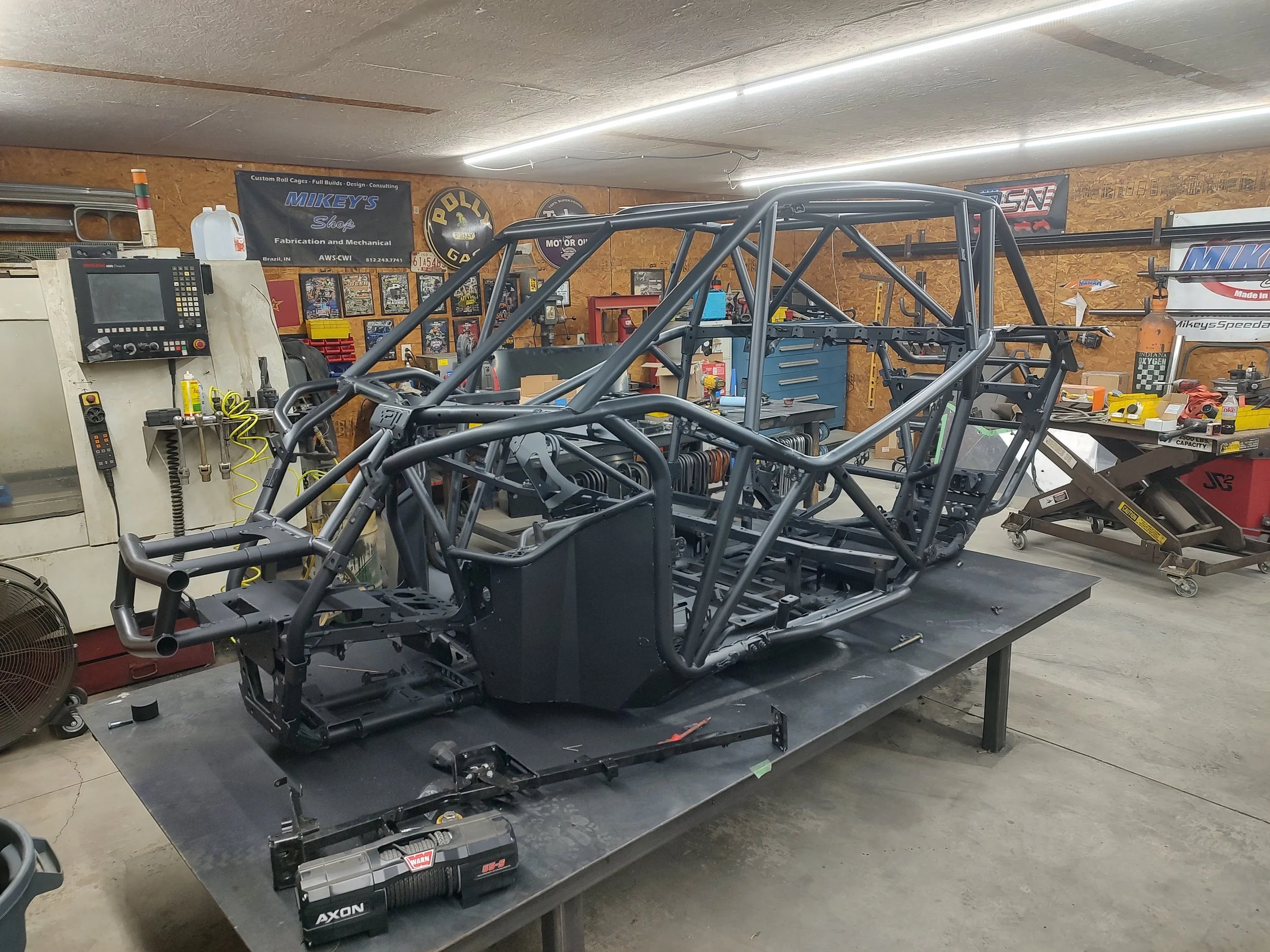 A partially assembled race car chassis in a workshop surrounded by tools, equipment, and posters on the walls.