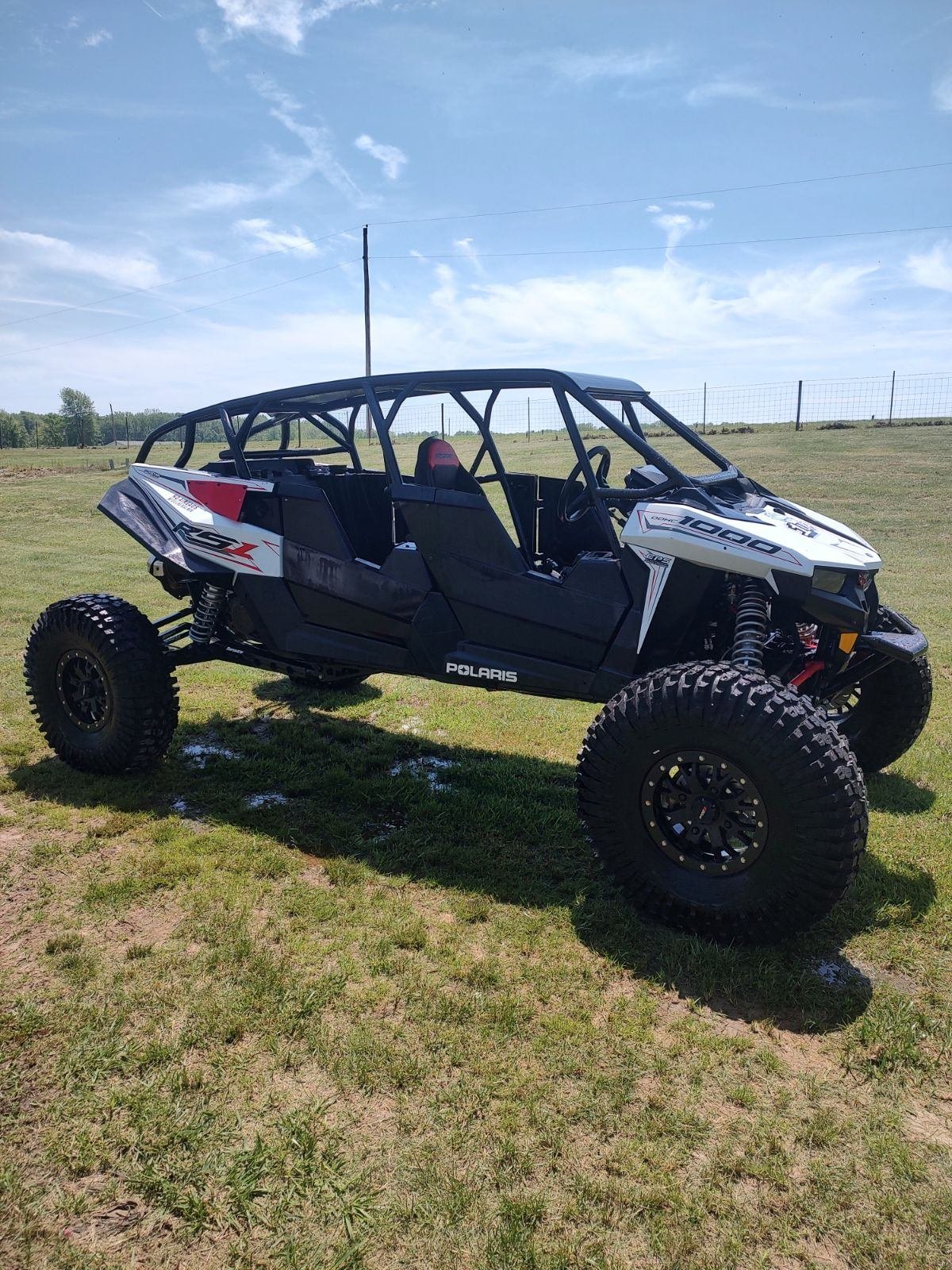A black Polaris RZR off-road vehicle with large tires, parked on grass field under a blue sky with some clouds.