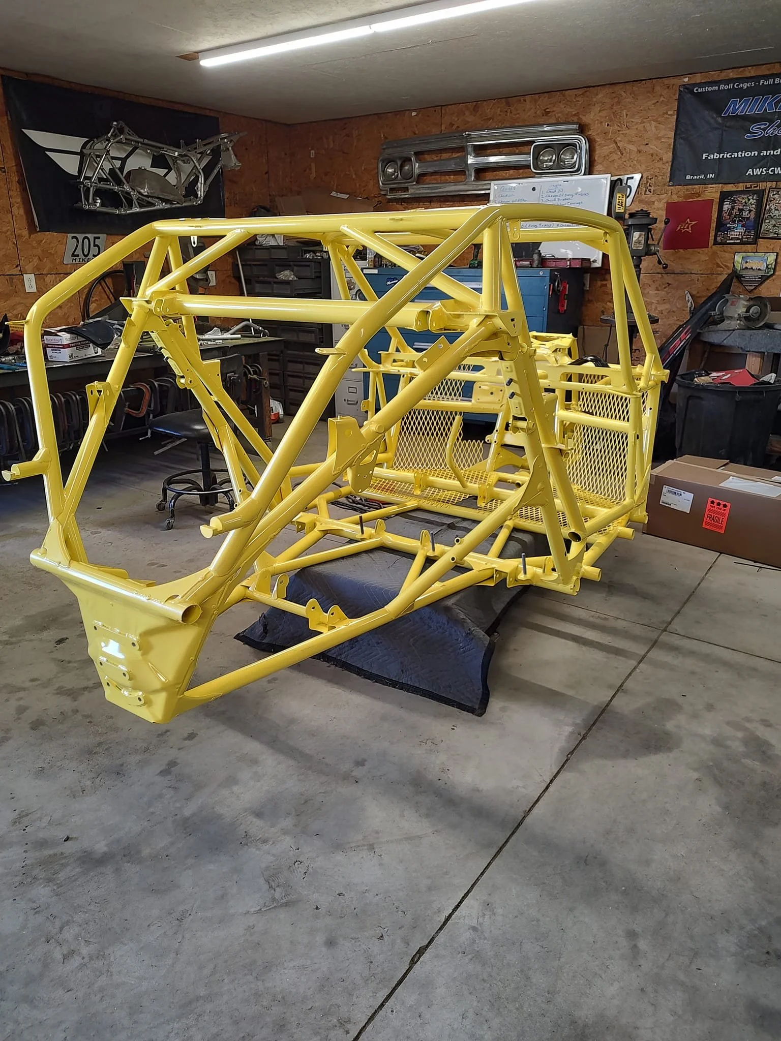 Yellow race car chassis in a workshop with various tools and car parts, including a headlight assembly and a metal frame, on the walls.