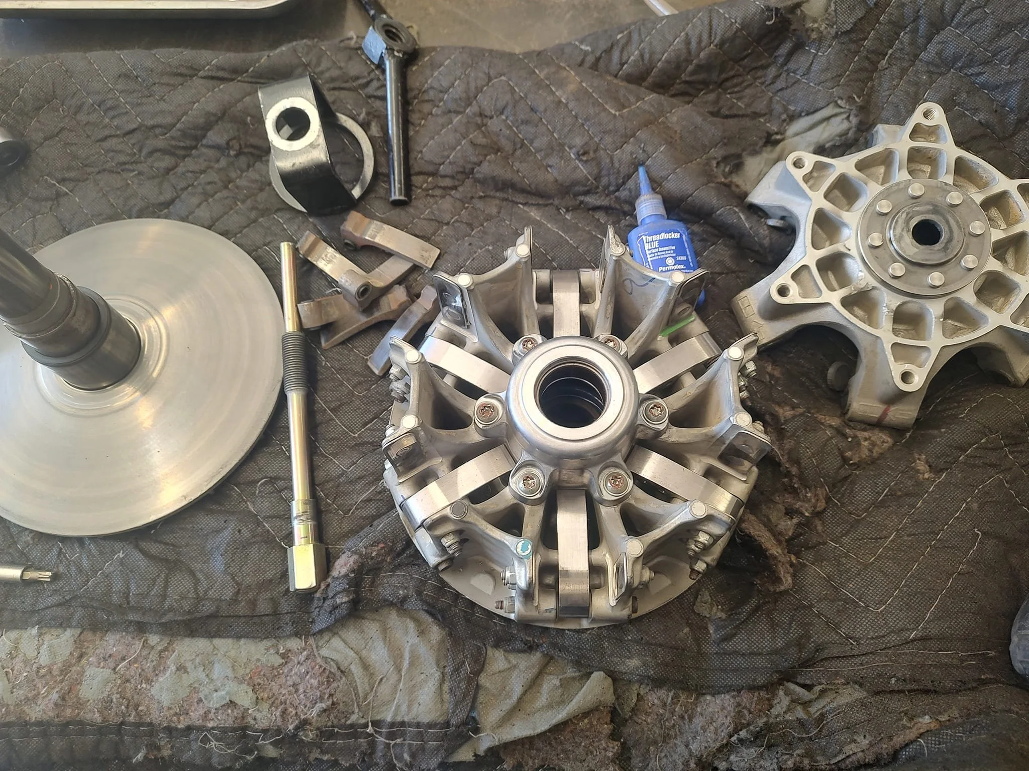 Disassembled motorcycle parts on a black padded surface, including a wheel hub, rotor, and various bolts and small metal components.
