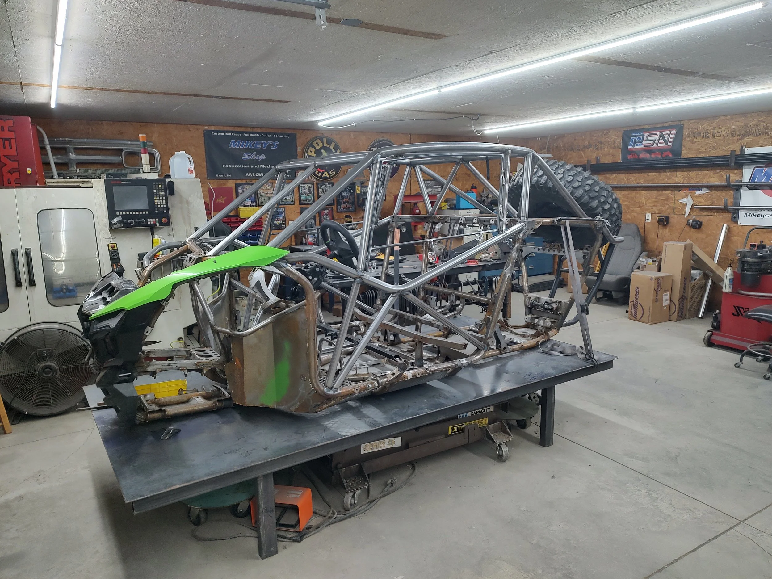A metal vehicle chassis or frame on a work table in a garage workshop, with a partial green body panel attached. The workshop has various tools, equipment, and posters on the walls, including a banner for Mikey's Shop and a sign for PSN.