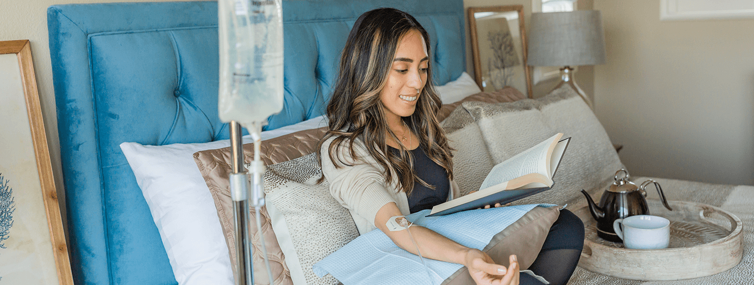 Woman sitting in bed with IV drip, reading a book, with teapot and cup on bedside tray.