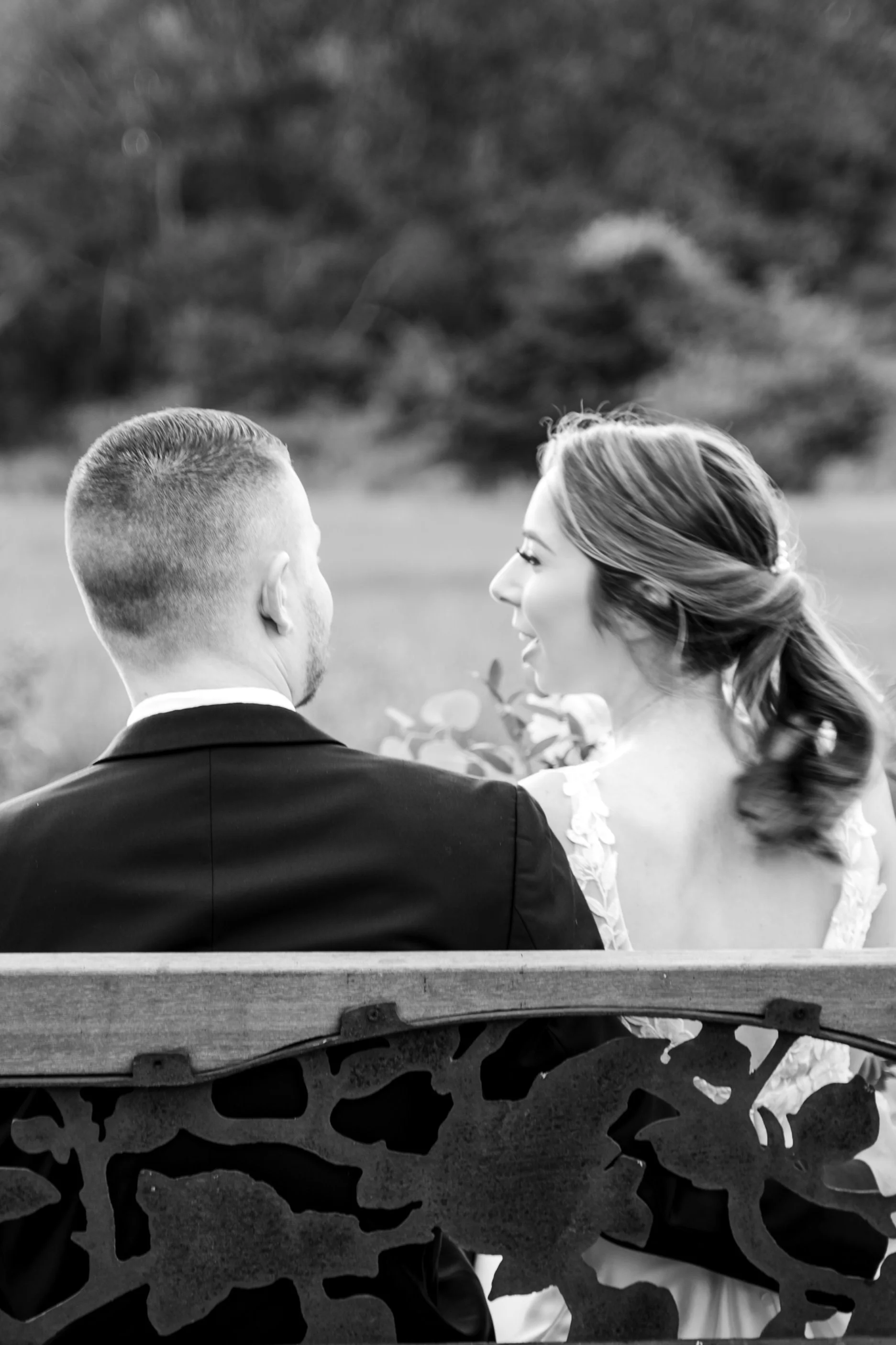 A black and white photo of a romantic moment between a man and woman sitting on a park bench, facing each other, with the woman smiling.