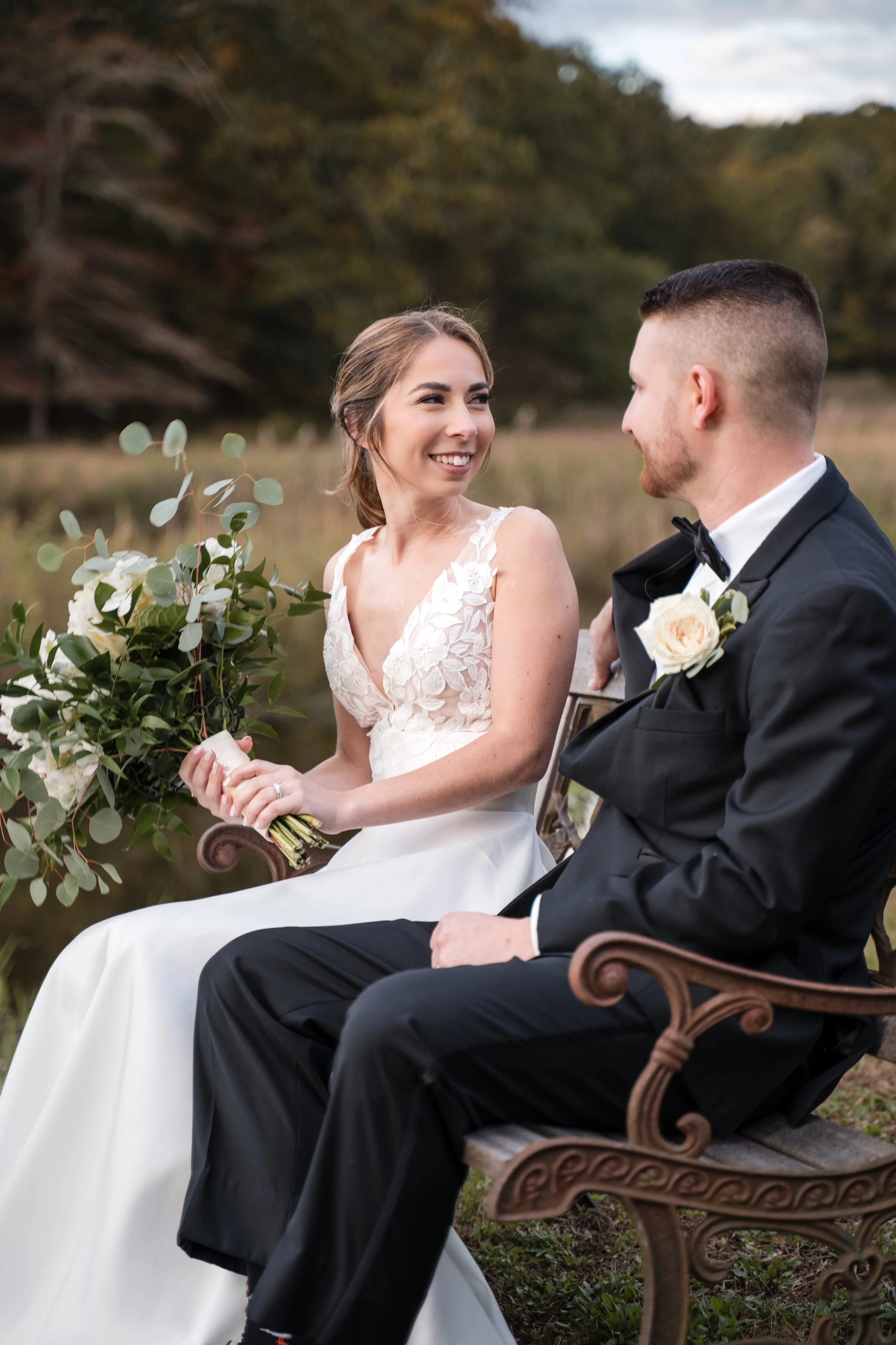 A bride and groom sit on a park bench outdoors, facing each other and smiling. The bride wears a white lace wedding dress and holds a bouquet of white roses and greenery. The groom wears a black tuxedo with a white shirt and black bow tie, with a whi