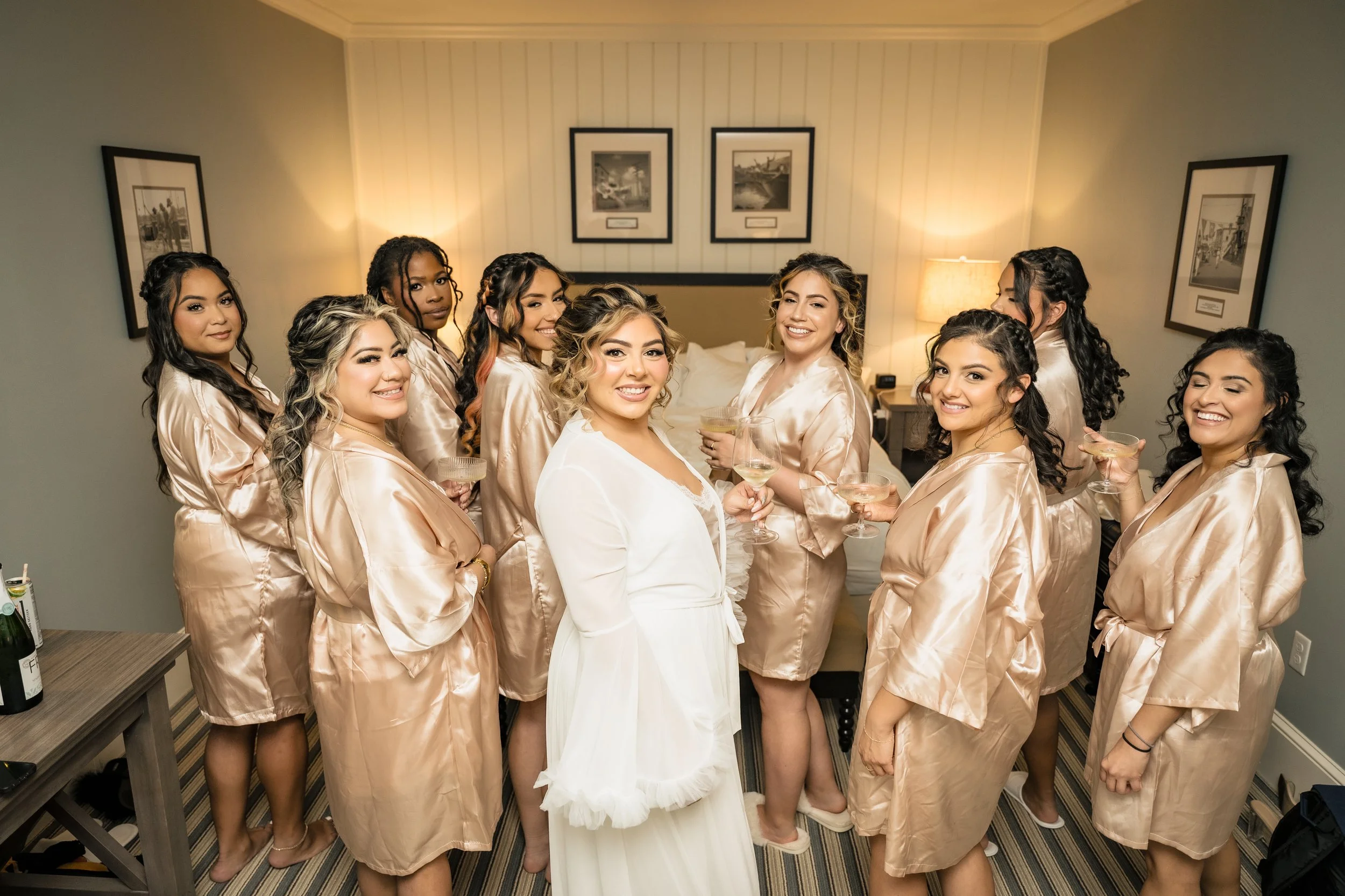 A bride in a white wedding dress stands in the center surrounded by nine women dressed in matching champagne-colored satin robes, all holding glasses of wine or champagne in a hotel room decorated with framed black-and-white photographs and soft ligh