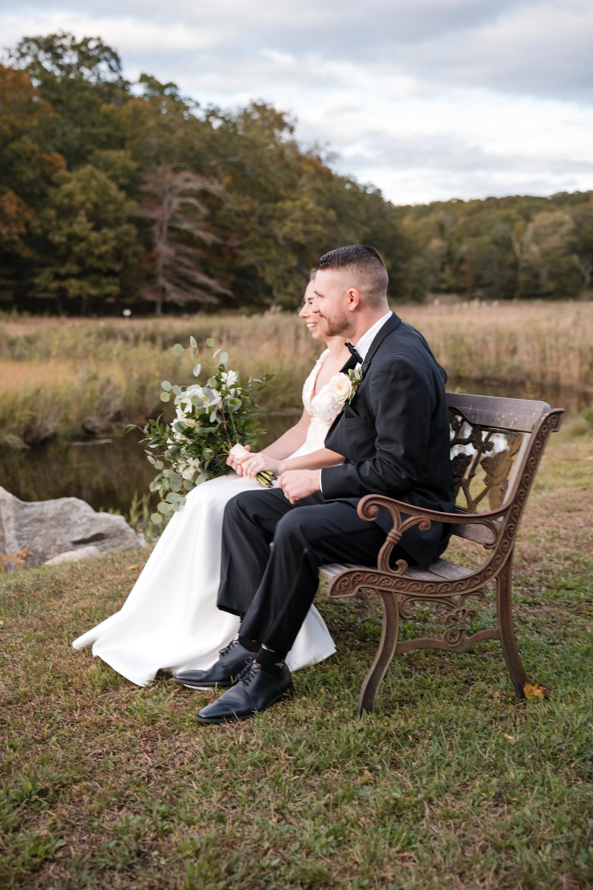 A bride and groom sitting on a bench outdoors near a pond, with trees in the background, dressed in wedding attire, smiling and holding hands.