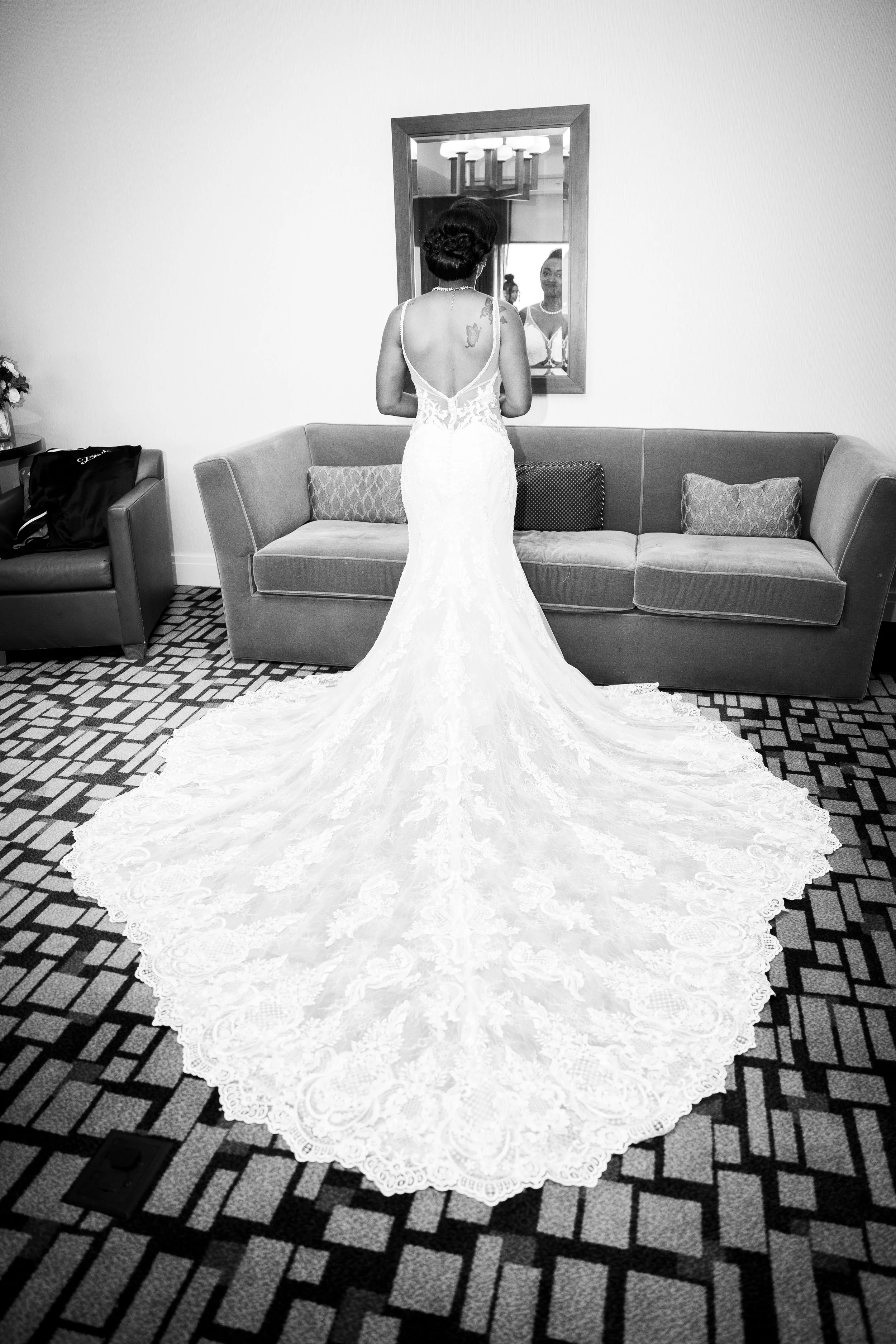 A bride in a white wedding gown with a long, lace train, standing with her back to the camera, looking into a mirror in a hotel room.