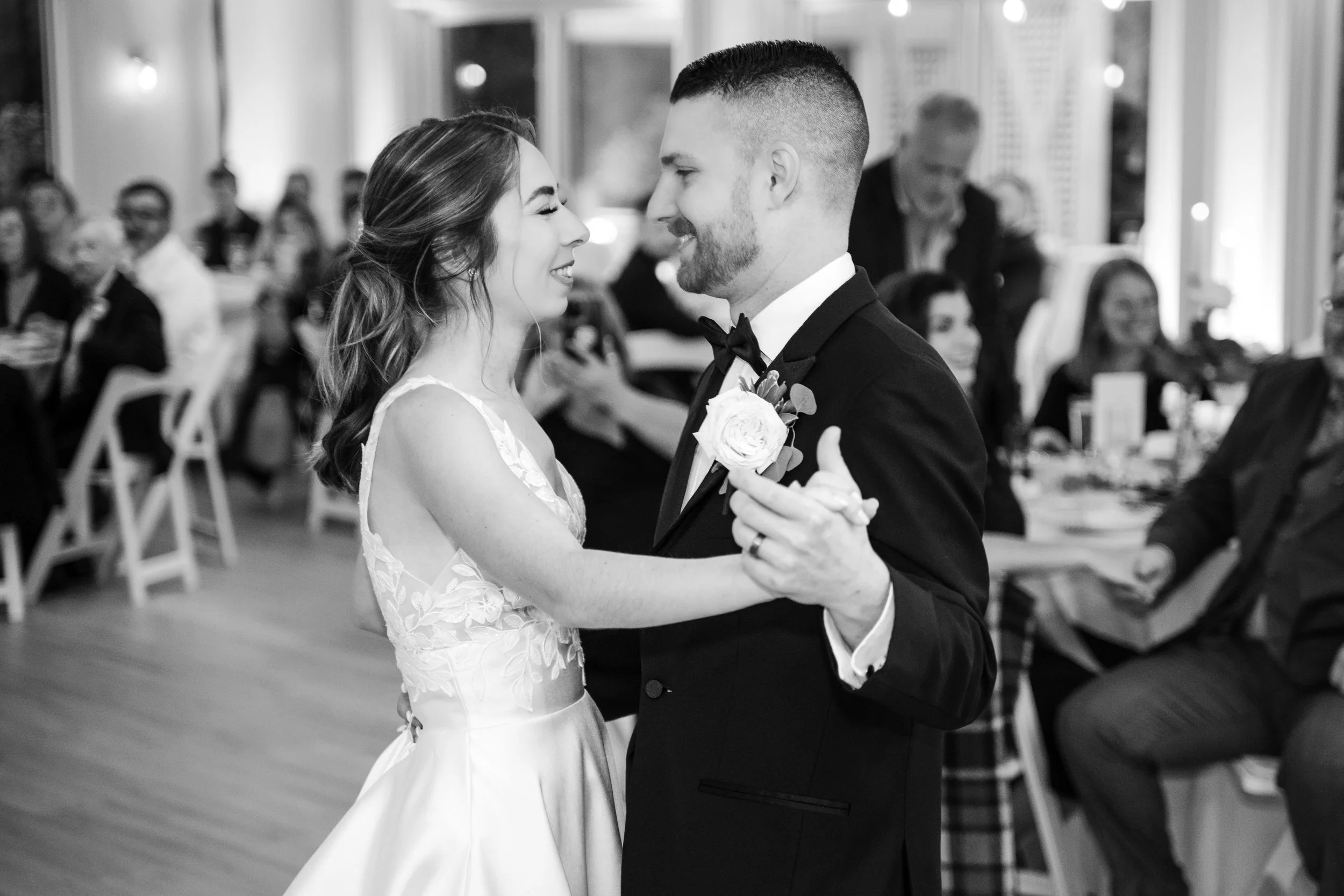 A bride and groom dancing at their wedding reception, smiling and looking into each other's eyes, with guests seated at tables in the background.