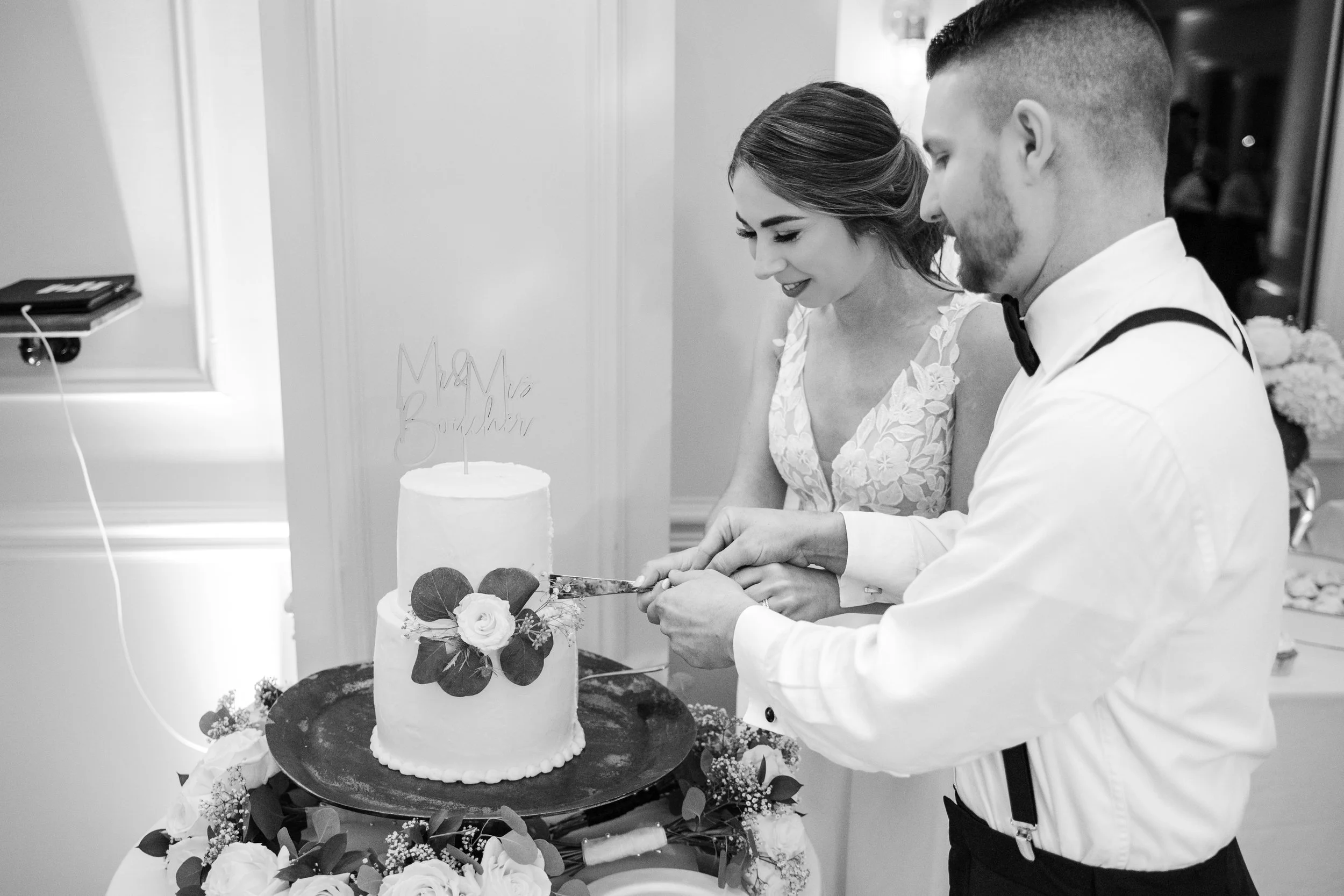 A black-and-white photo of a bride and groom cutting a wedding cake together. The bride is smiling and looking down at the cake, with her hair styled in a side part and wearing a floral lace dress. The groom is dressed in a white shirt with suspender