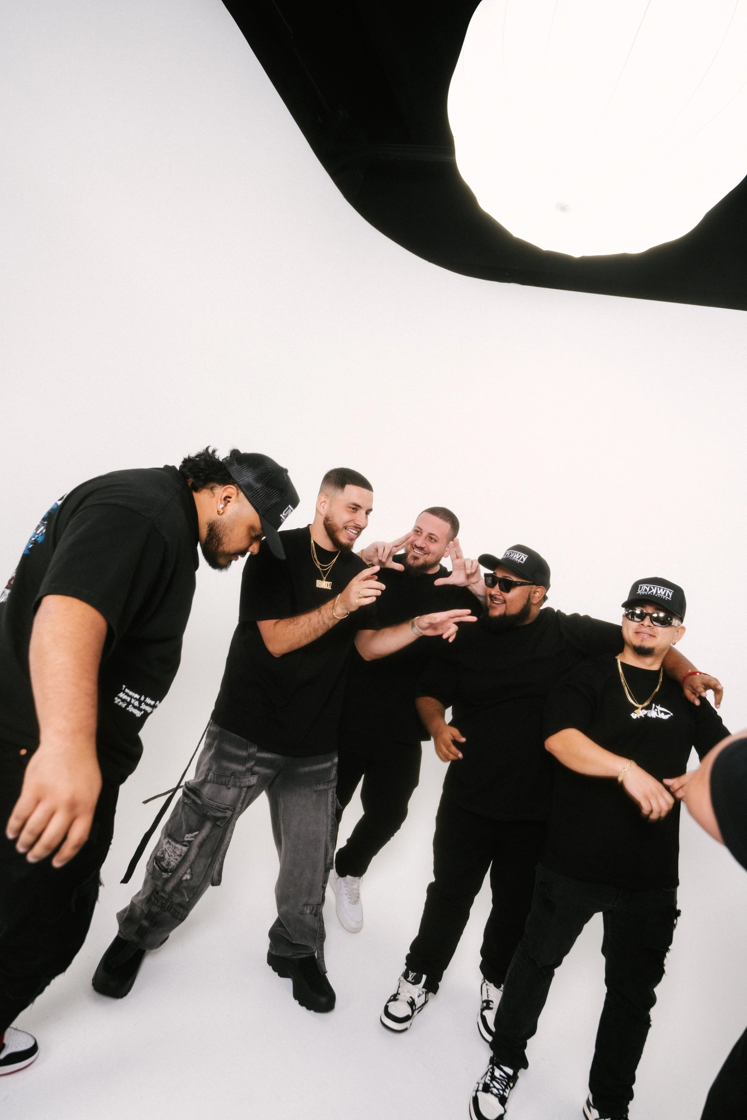 Group of five men in black outfits posing and having fun in a photo studio with a white background, taken from a low angle with a large roof light overhead.
