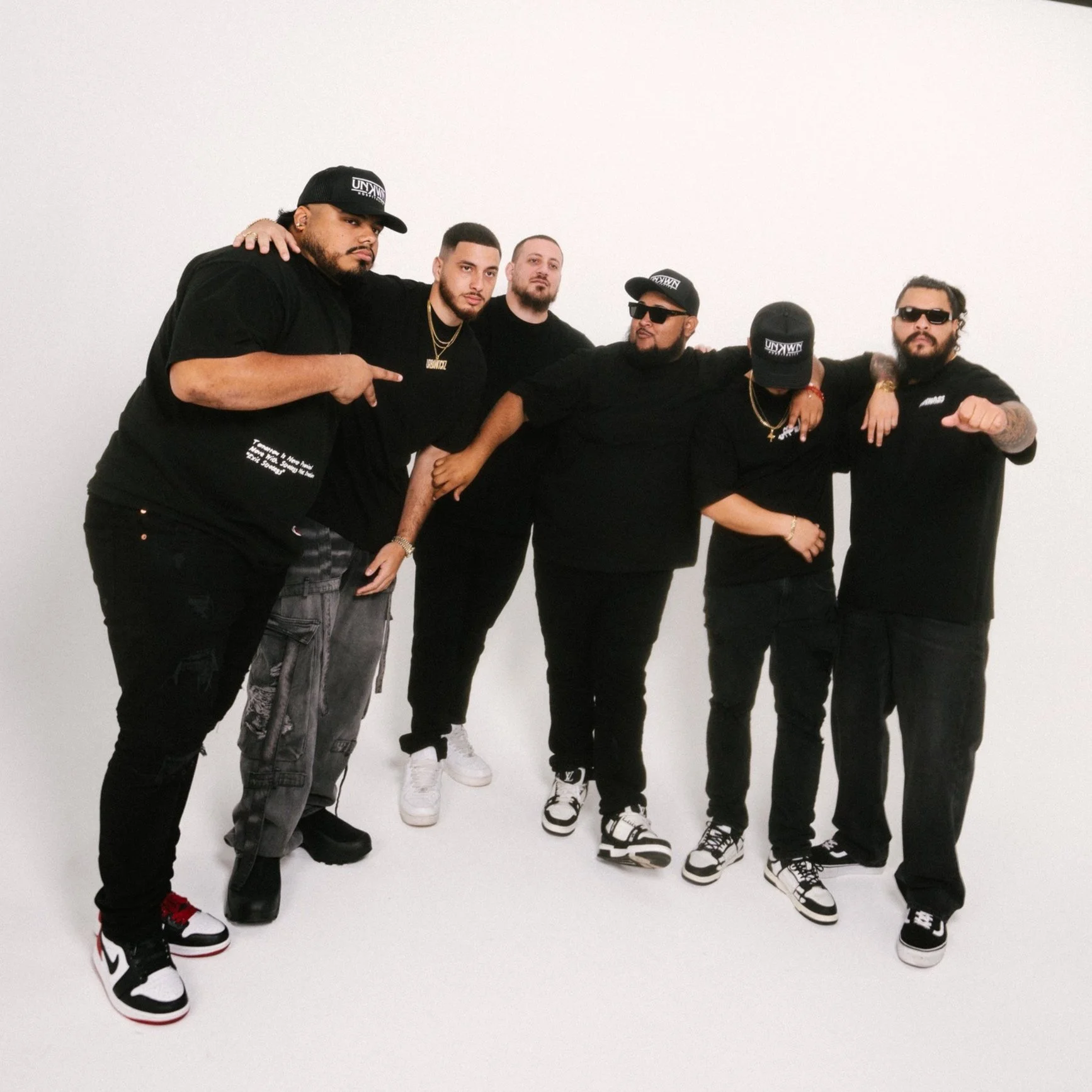Six men dressed in black outfits posing against a plain white background, with some wearing sunglasses and caps, and displaying hand gestures and arm placements.