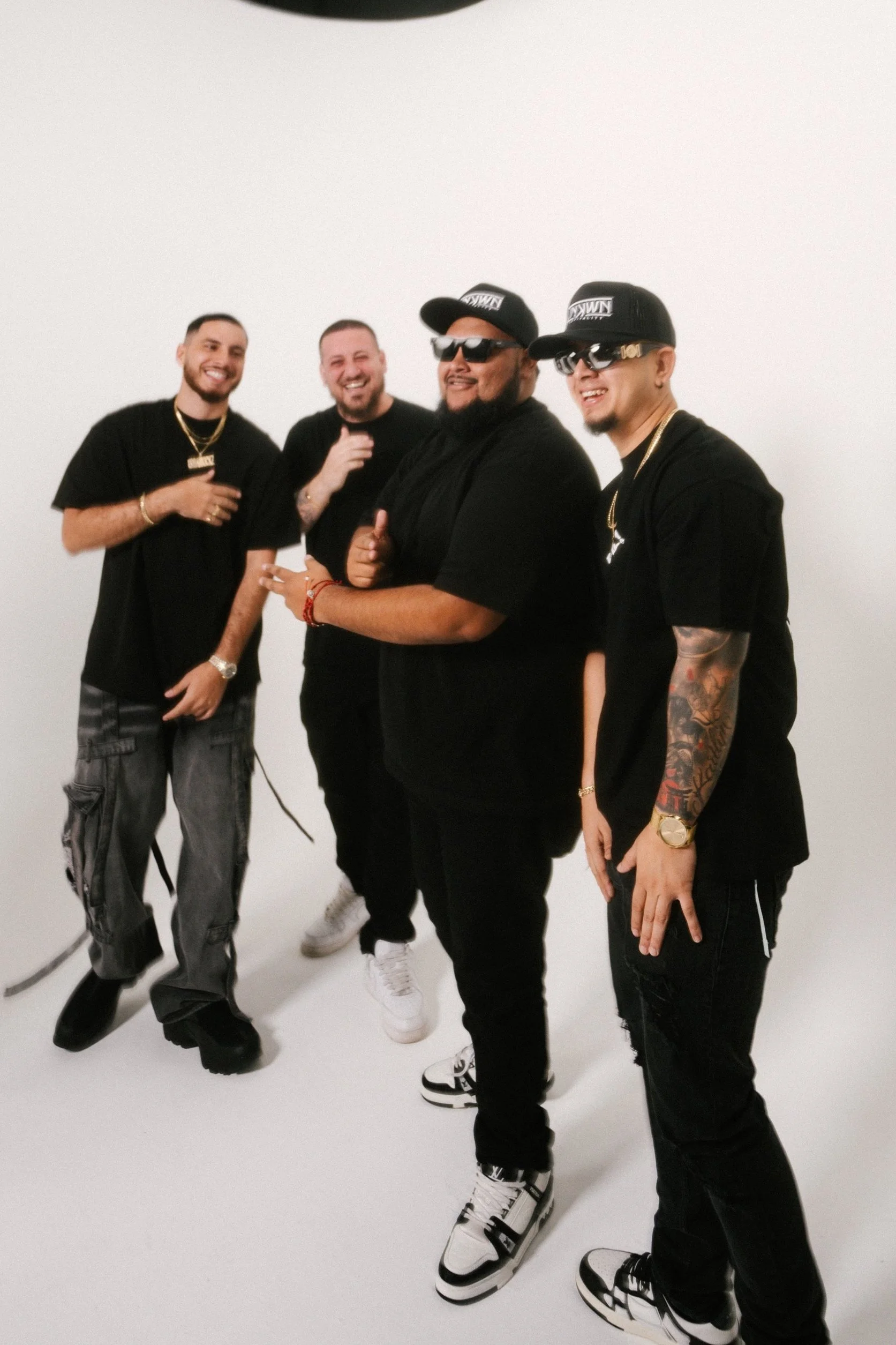 Four men standing together, smiling, in a white studio. They are dressed in black clothing with accessories, and three are wearing sunglasses and caps. The group appears relaxed and happy.