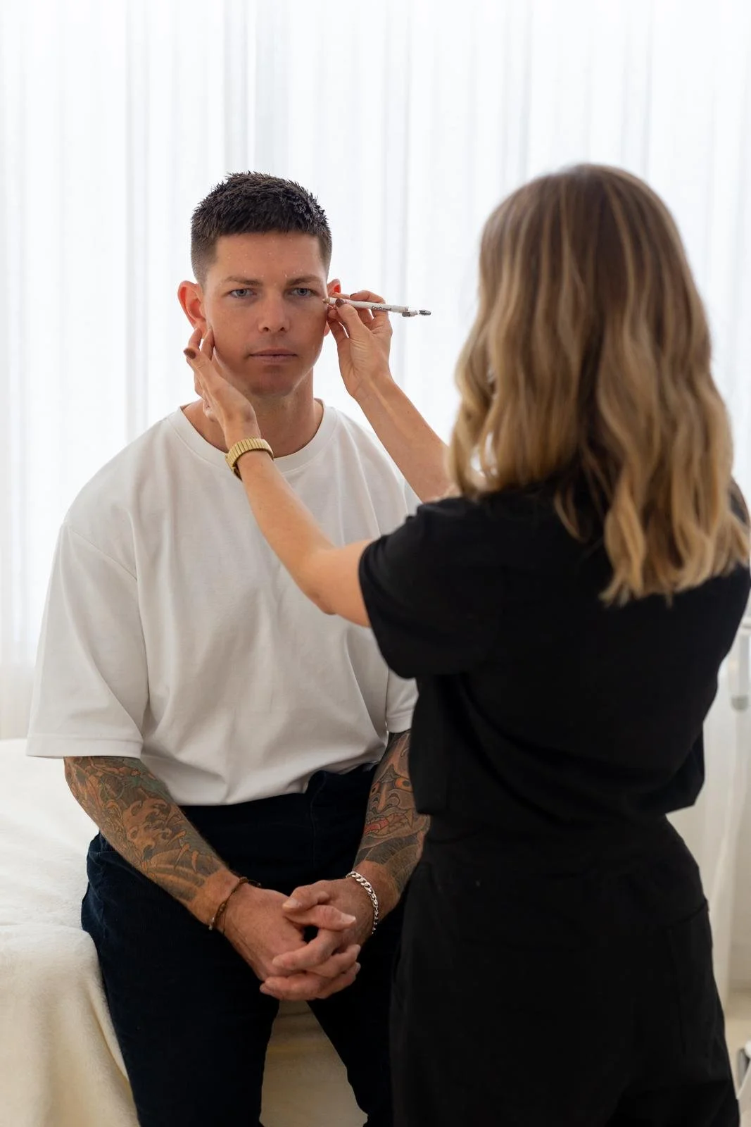 A woman is applying makeup or performing a cosmetic procedure on a man sitting on a bed in a well-lit room with white curtains.