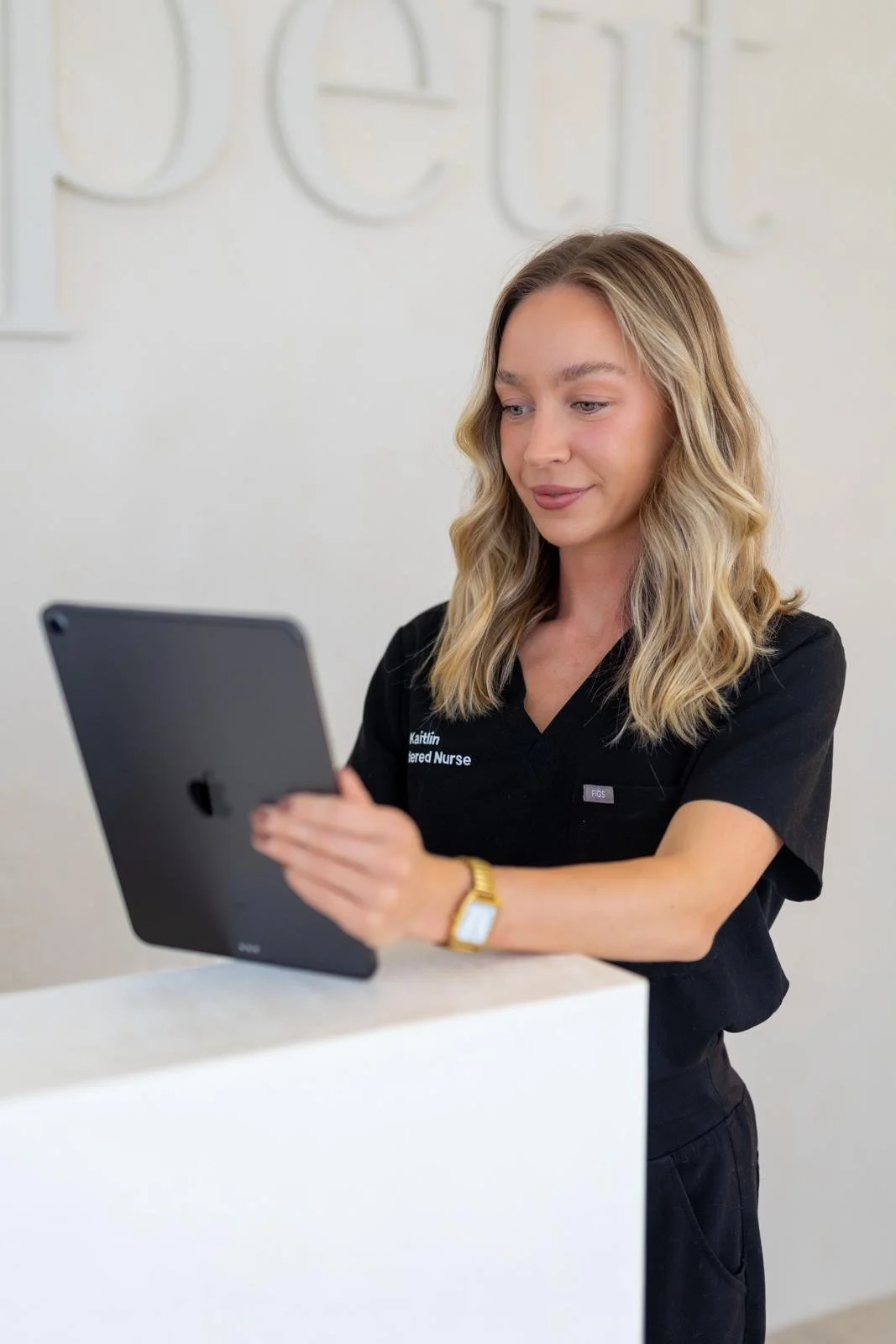 A woman in a black uniform standing at a white counter, holding an iPad, with a neutral background and partial text on the wall behind her.