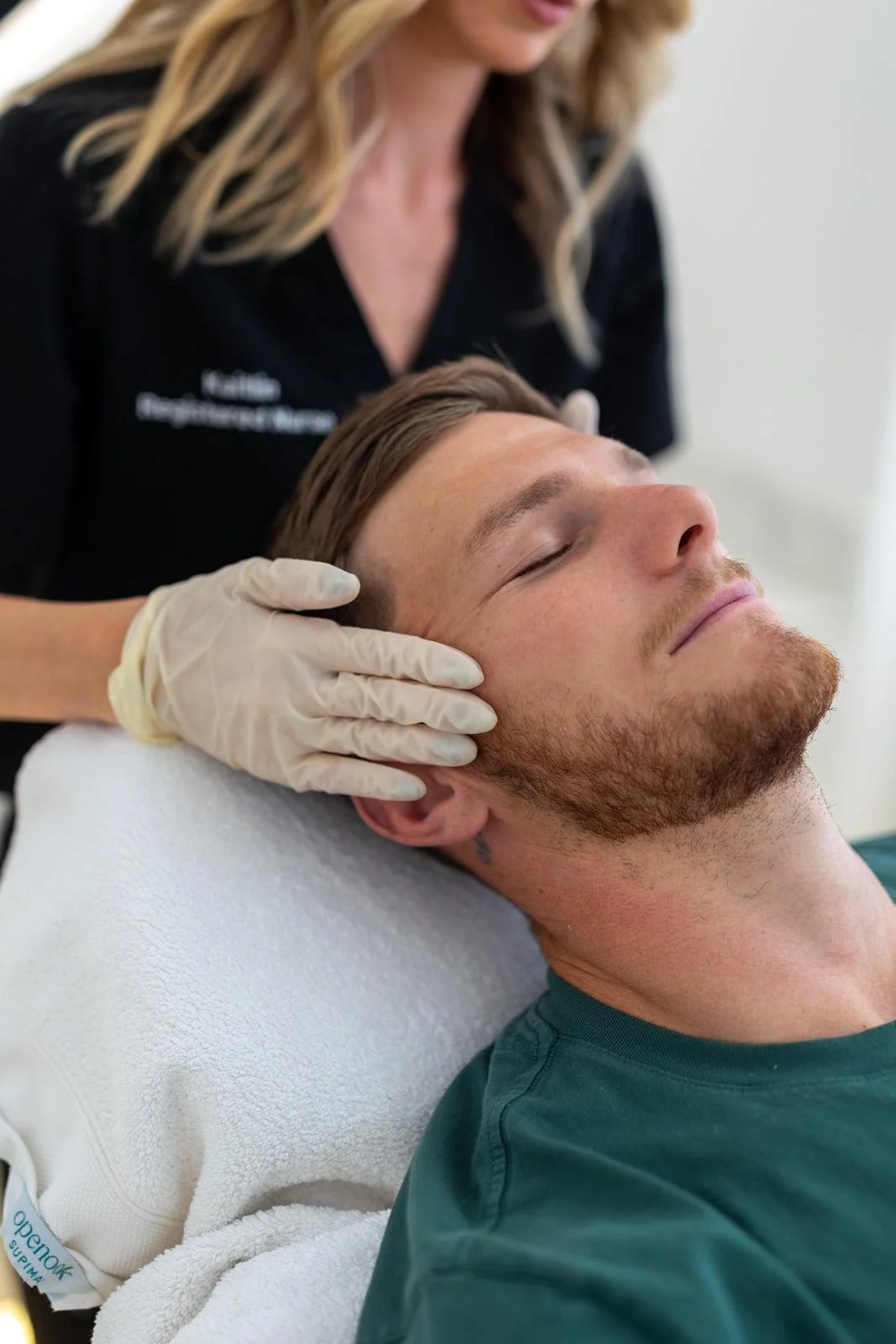 A man is lying on a treatment bed with his eyes closed, receiving a facial or skincare treatment from a professional wearing gloves.