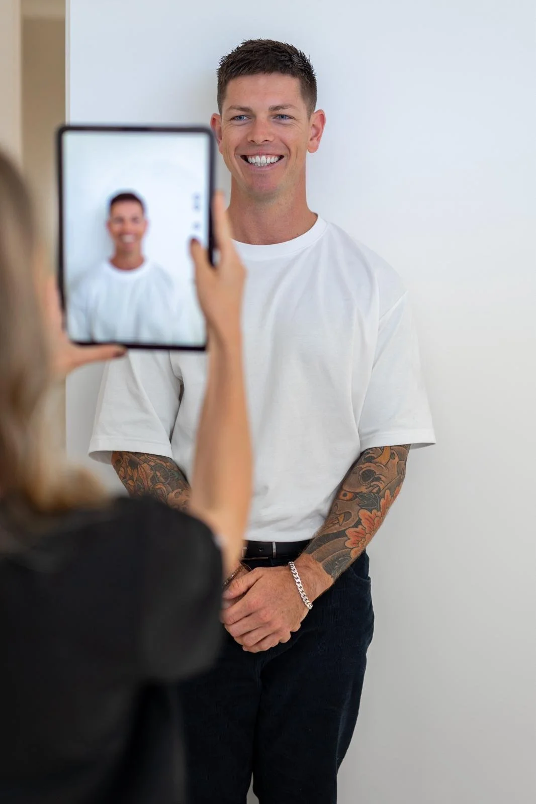 A man with short brown hair, wearing a white t-shirt and tattoos on his arms, smiling as a woman takes his photo with a tablet.