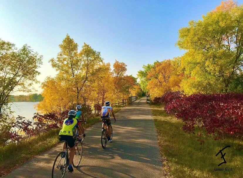 Two cyclists riding the paved Central Lakes Trail beside the water, gold trees overhead and red sumac along the edge