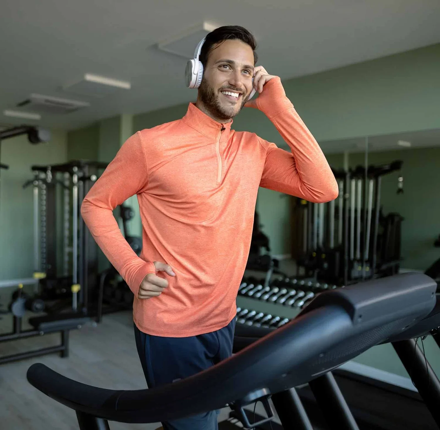 A man in orange workout shirt running on a treadmill in a gym, smiling and listening to music with headphones.