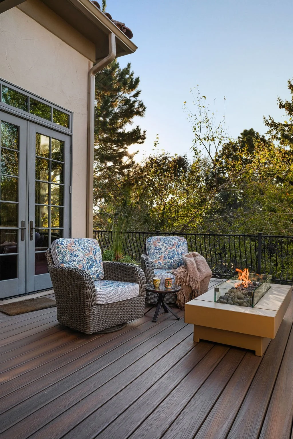 Outdoor deck with wicker chairs, patterned cushions, throw blanket, small table, and lit fire pit overlooking trees