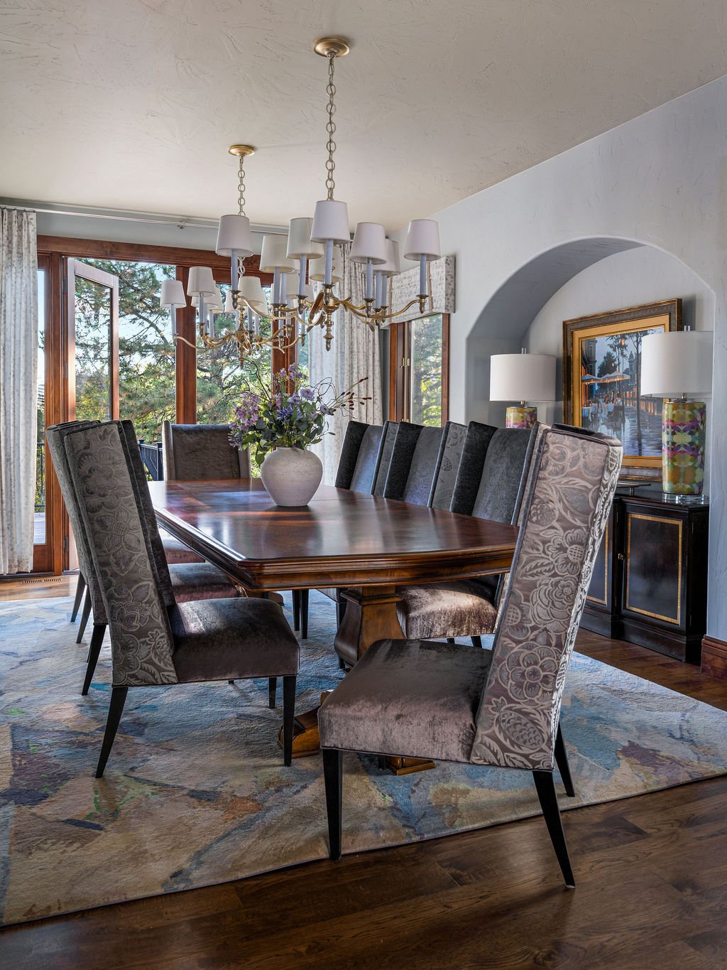 Elegant dining room with a large wooden table, upholstered chairs, twin chandeliers, floral centerpiece, and windows overlooking trees